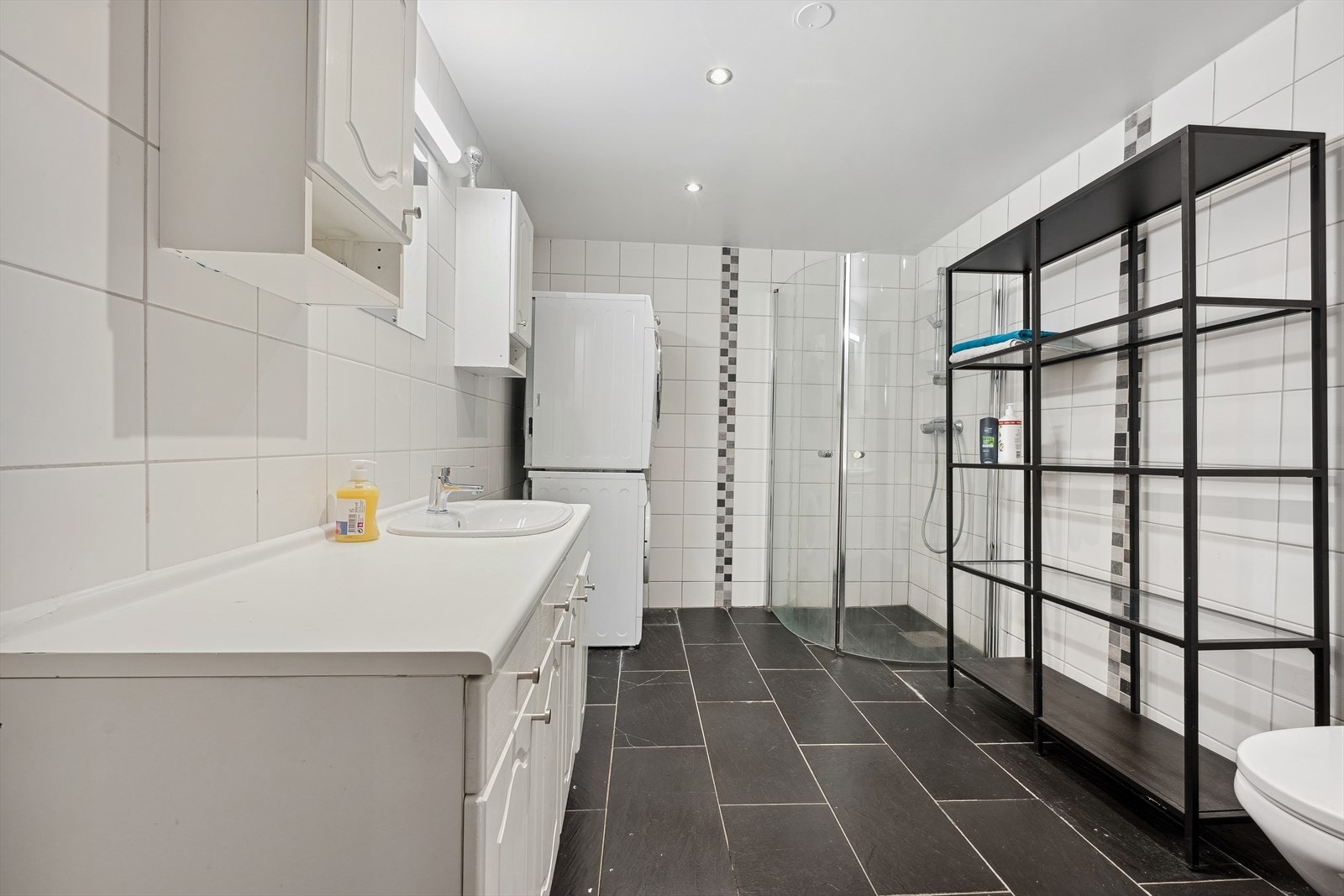 Helfliset baderom med nedfelt servant på innholdsrik skapinnredning med hvite fronter, overskap, speil med lys, toalett og dusjhjørne med glassvegger. Galleribilde