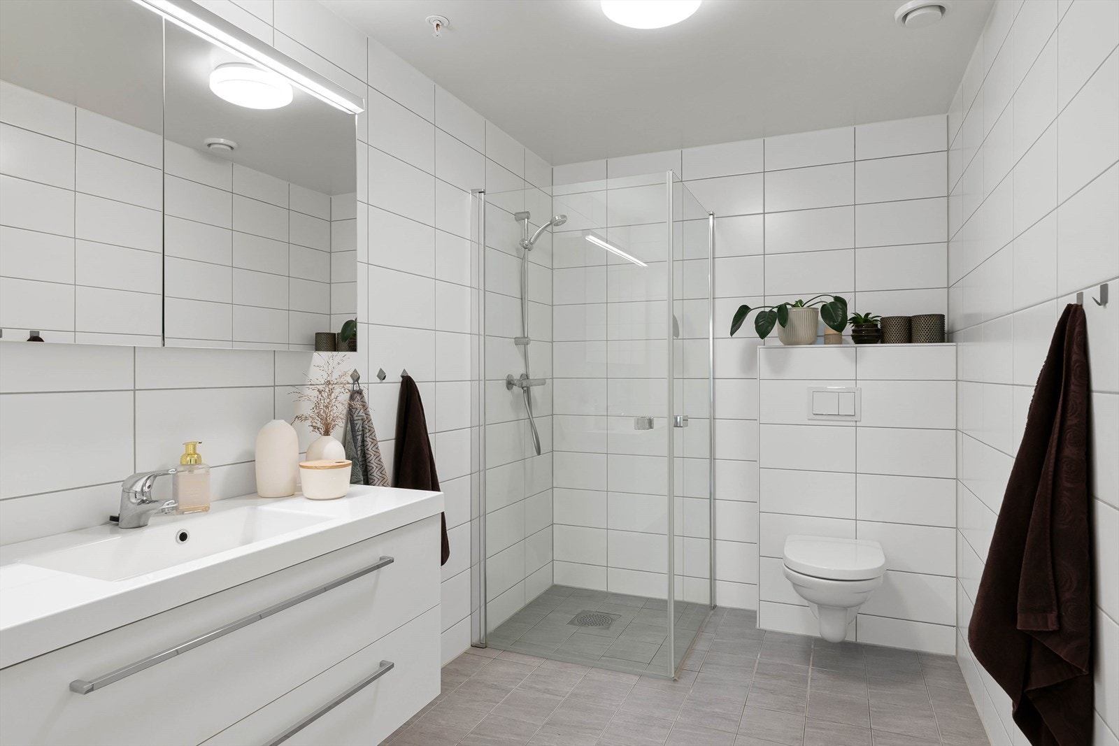 Lekkert bad med servant i innredning med skuffer, speilskap med lys over vask, dusjvegger som kan slås inn og vegghengt toalett. Galleribilde