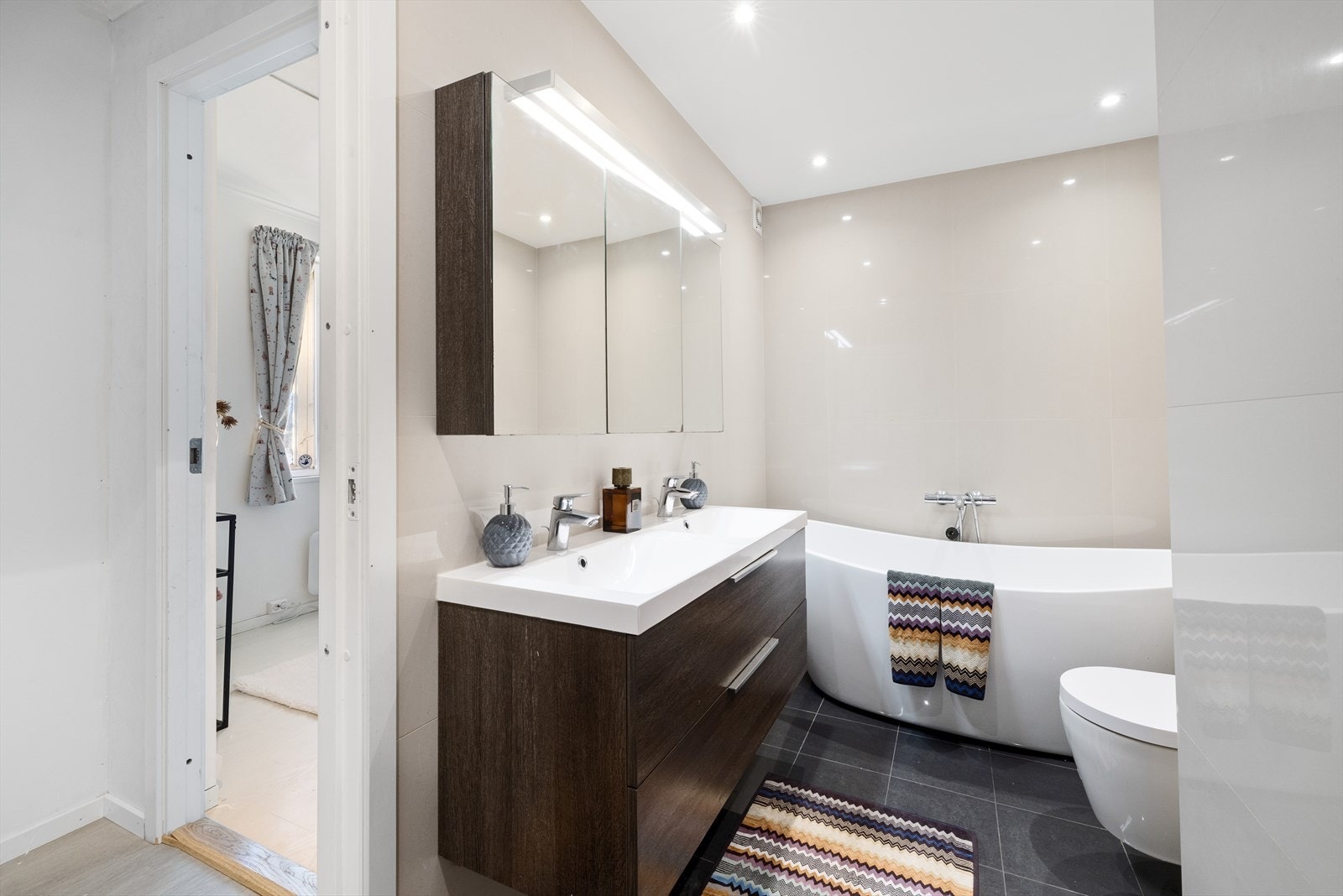 Lekkert, flislagt bad som er innredet med veggmontert toalett, dusjnisje med rainshower, badekar, dobbelservant og skapspeil med lys. Galleribilde