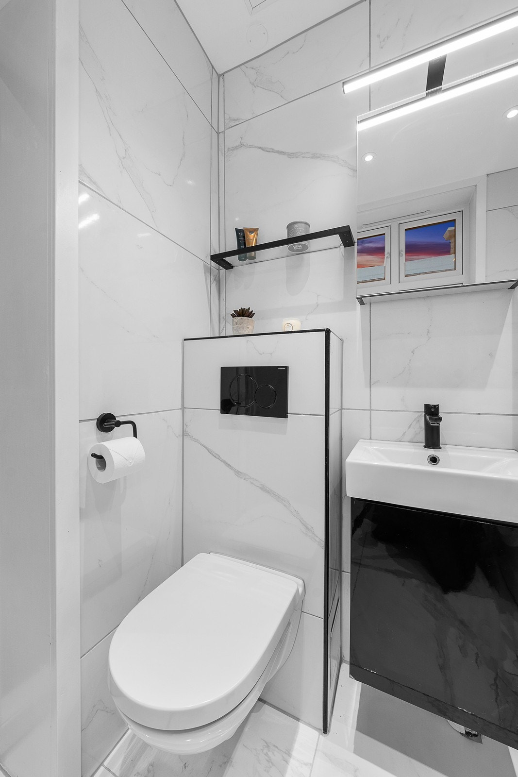 Badet består av dusjhjørne med svingbare dusjdører i herdet glass, vegghengt toalett, heldekkende servant med underskap og speilskap med belysning over. Galleribilde