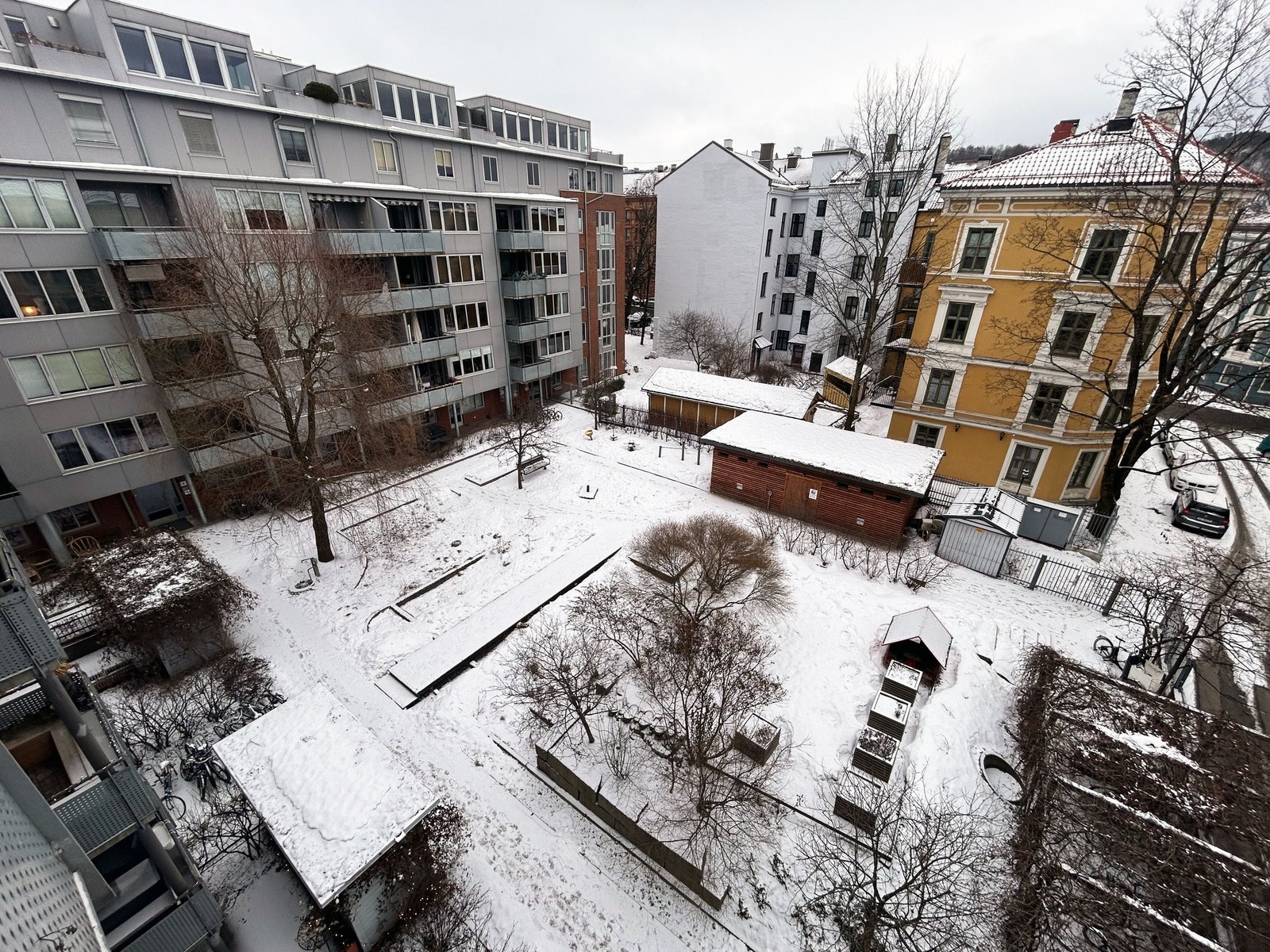 Utsikt fra balkongen ned på felles inngjerdet hageanlegg med sitteplasser, dyrkningskasser og lekeapparater Galleribilde