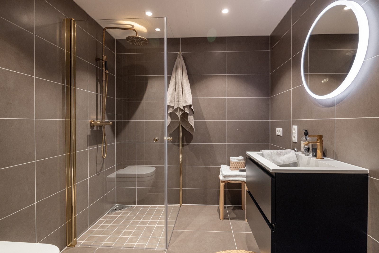 Baderommet har mørke fliser på gulv og vegger som gir en behagelig atmosfære, og skaper et lunt og harmonisk uttrykk. Det er vannbåren gulvvarme, downlights i himlingen og mekanisk avtrekk via balansert ventilasjon. Galleribilde