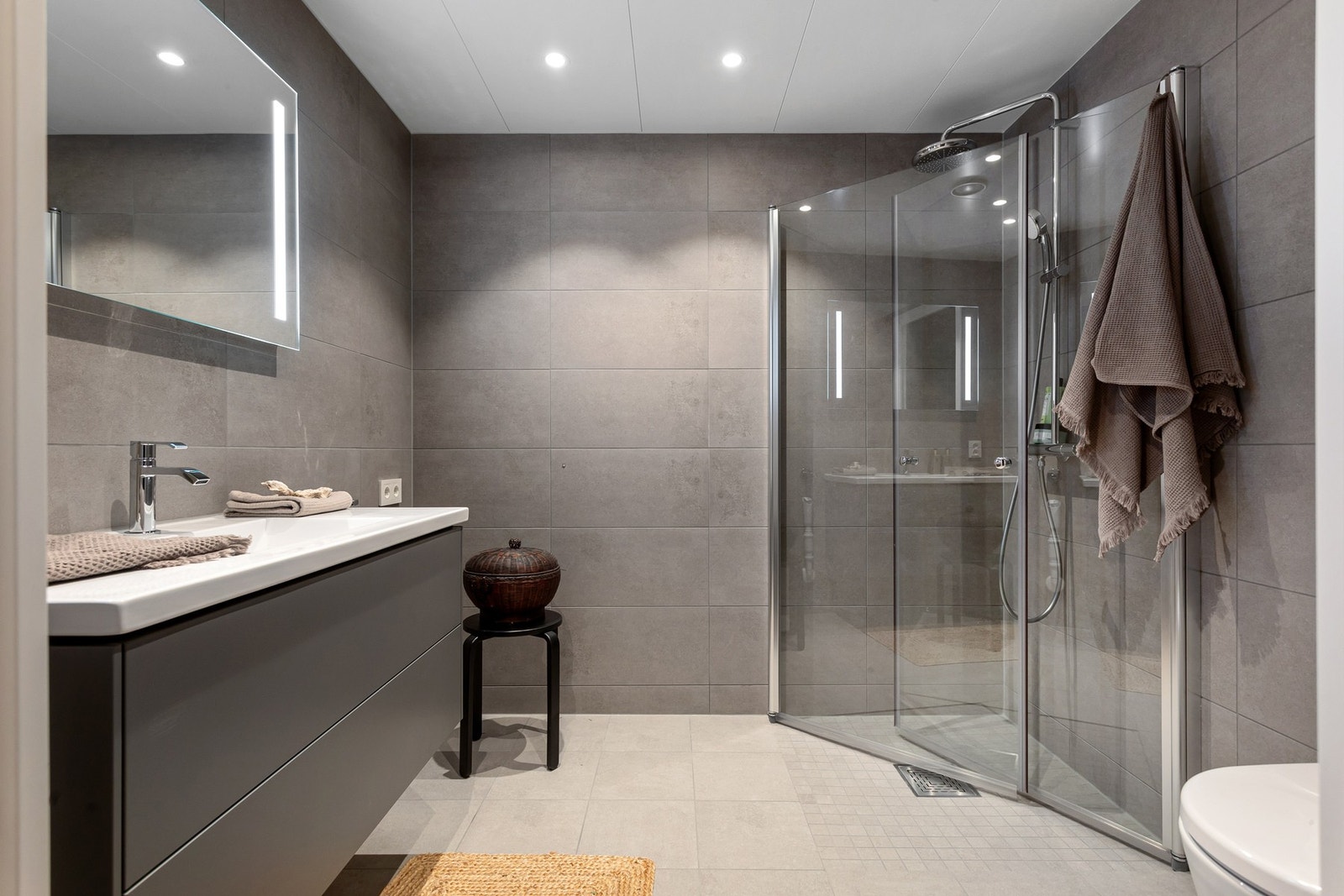 Baderommet har matte porselenfliser fra Vitra med et stilrent design, deilig gulvvarme og LED-downlights i himlingen. Flisene har fint spill i seg som tilfører liv til det grå og passer godt sammen med armatur og detaljer i krom. Galleribilde