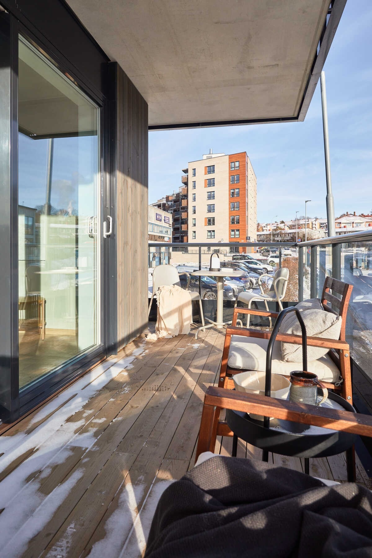 Balkongen er overbygget, har terrassebord på gulv og rekkverk av metall og glass. Det er også montert utebelysning og stikkontakt på vegg. Galleribilde