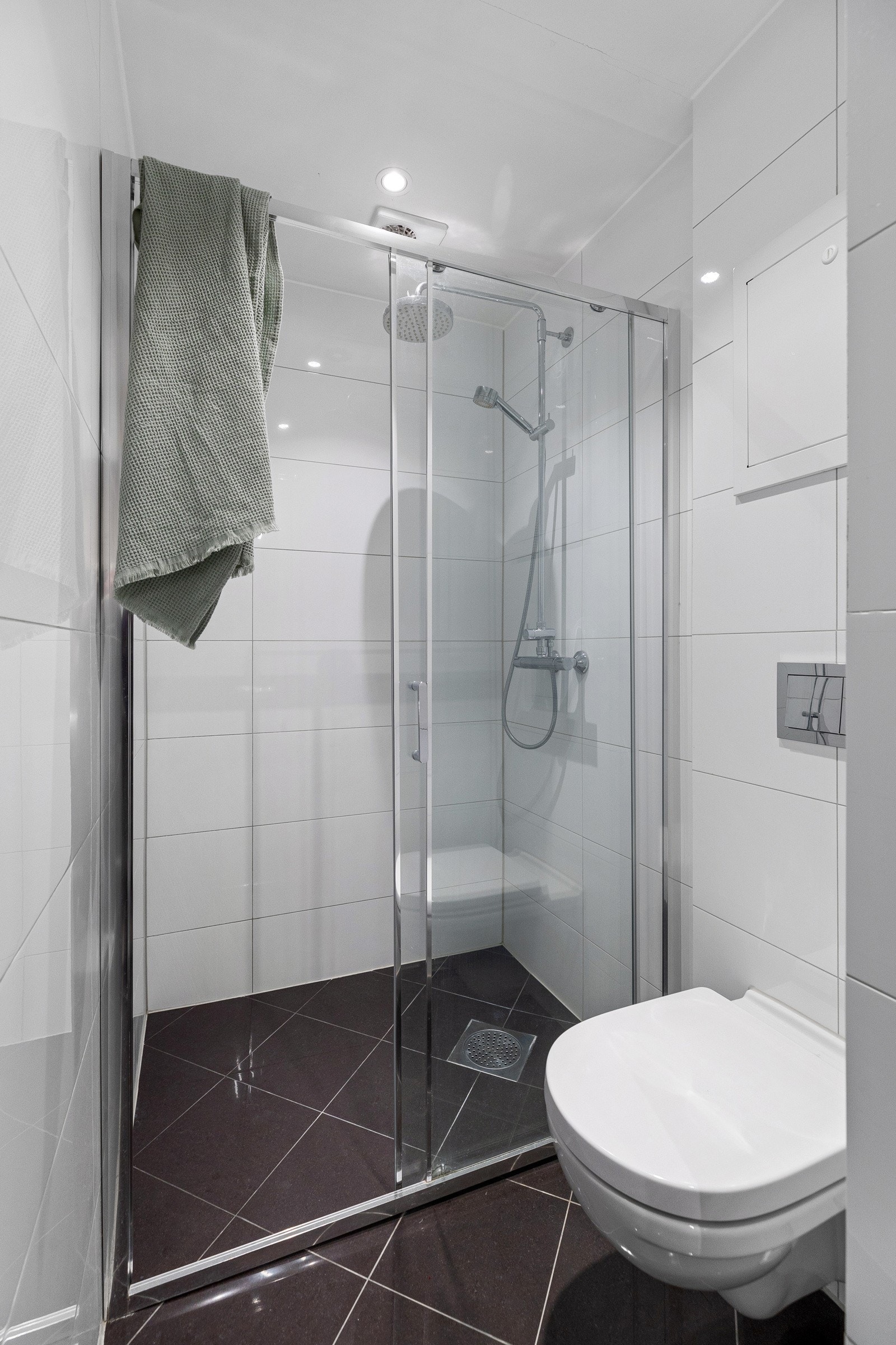 Badet inneholder servantskap med speilskap og lysarmatur, vegghengt toalett og et dusjhjørne med skyvedør og regnfall. Galleribilde