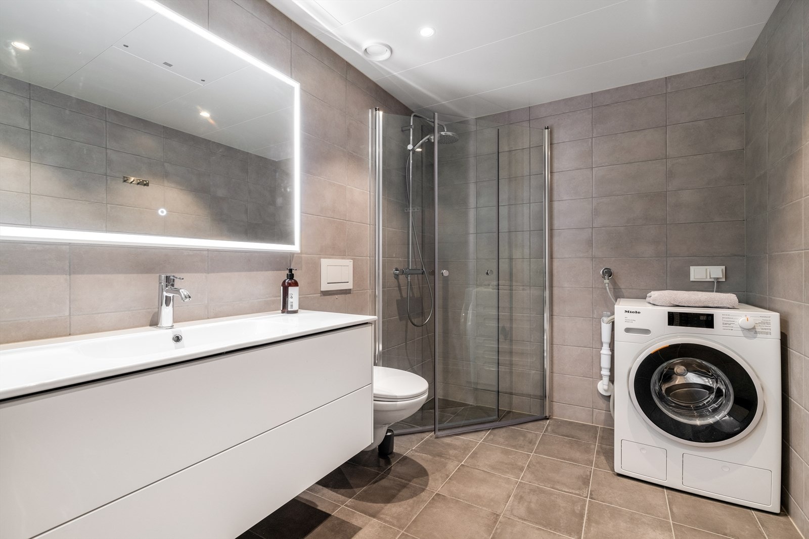 Det andre badet har inngang fra entreen, og har flis på gulv og vegger. Her er det vegghengt toalett, dusjhjørne med vegger, innredning og opplegg for vaskemaskin. Det er varmekabler i gulv. Galleribilde