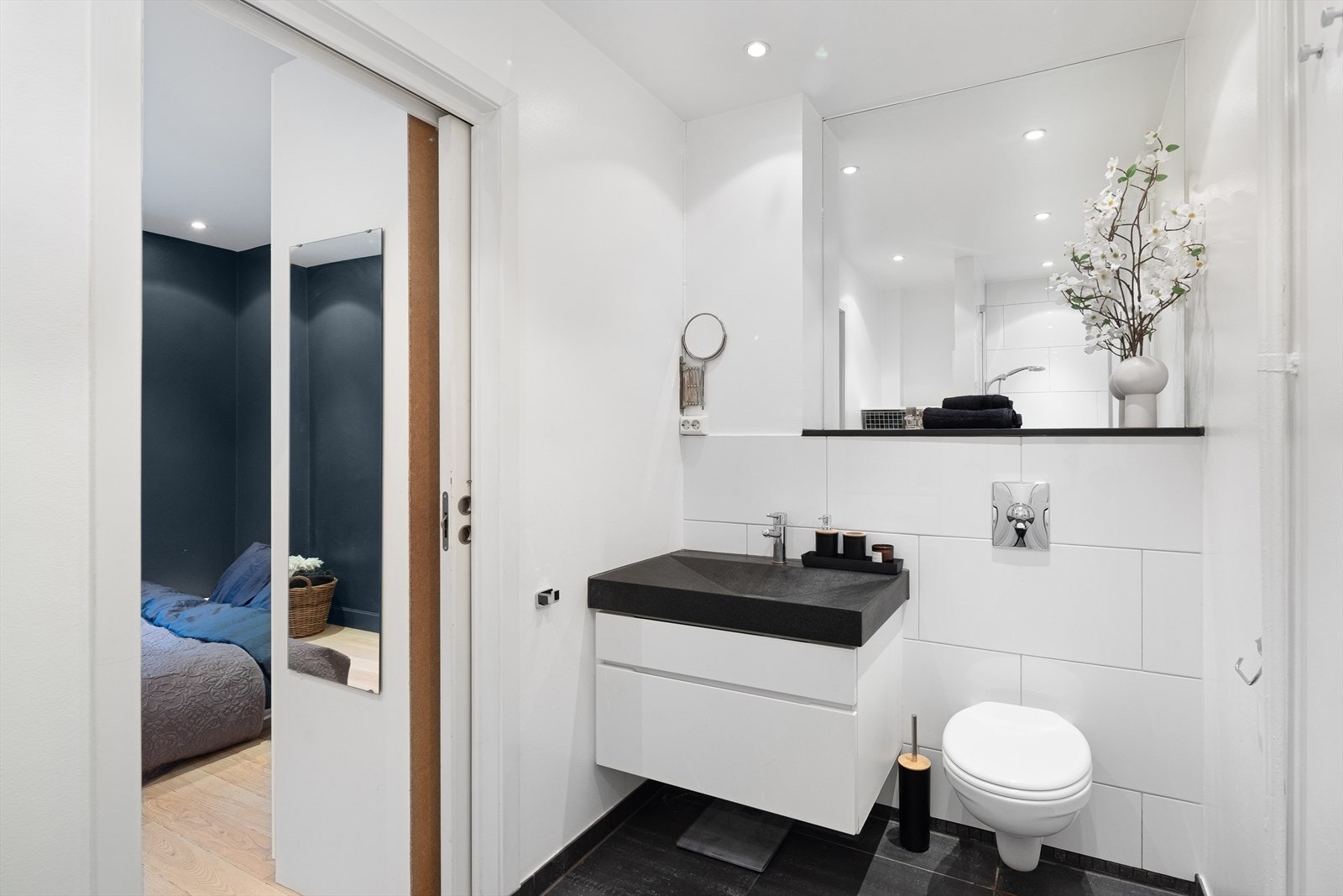 Badet er utstyrt med nedsenket dusjnisje og dør i herdet glass, helservant med underskuffer, speilfelt over servant, vegghengt toalett og hyller over nisje for opplegg til vaskemaskin. Galleribilde