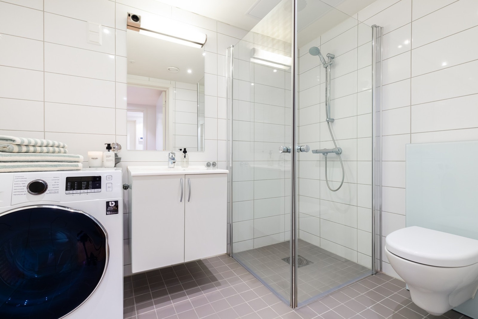 Boligens bad er bygget opp med grå fliser på gulv og lyse fliser på vegger. Det er downlights i himling og varme i gulv. Innredningen består av servant på underskap, speil med belysning og vegghengt toalett. Galleribilde