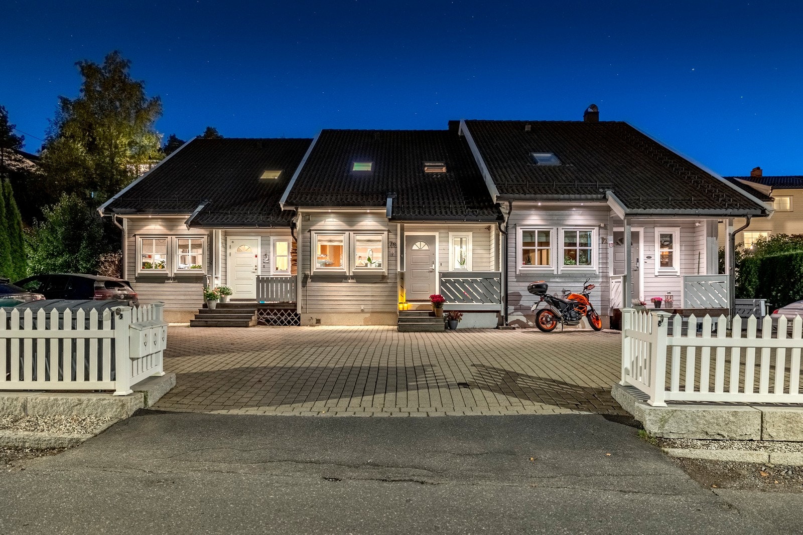 Foran rekken med boliger er det en felles, steinbelagt gårdsplass, og ut mot veien er det satt opp hvitmalt gjerde. Galleribilde