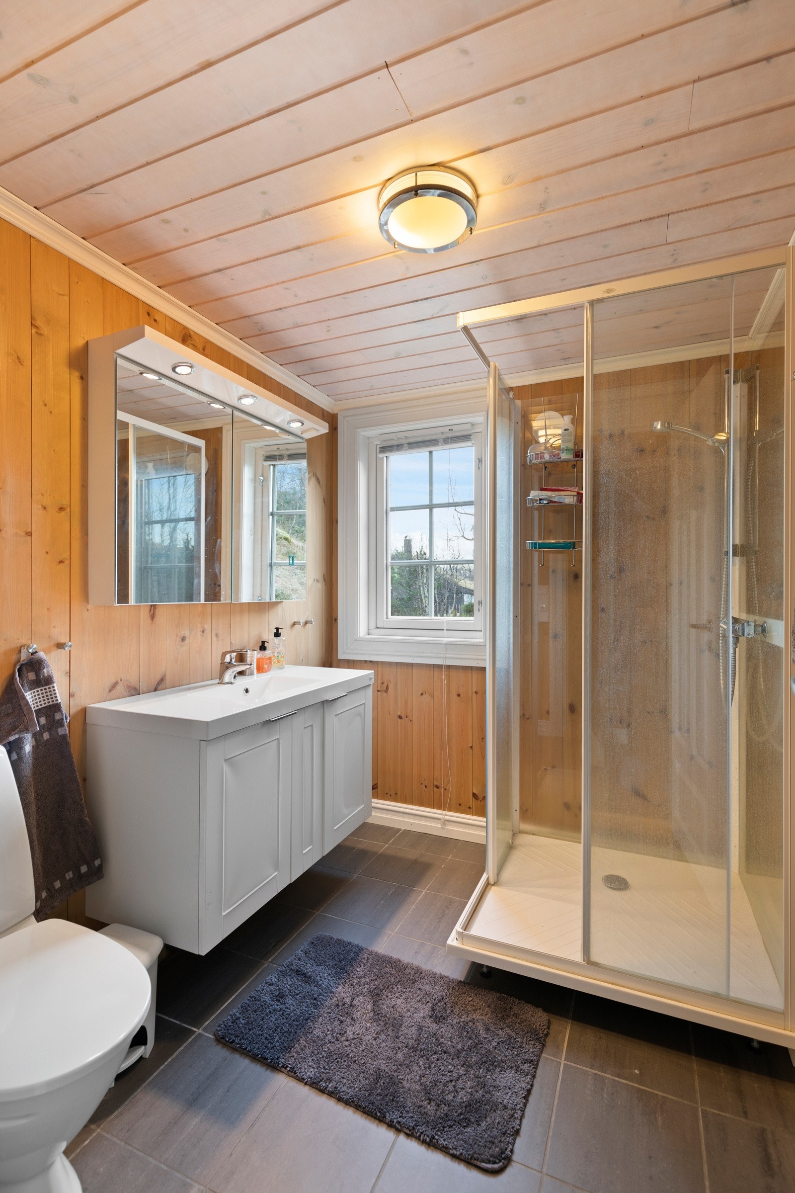 Badet har flis på gulv og panel på vegger. Rommet er utstyrt med varmekabler i gulv, dusjkabinett, toalett og baderomsinnredning med servant, speilskap og belysning. Galleribilde