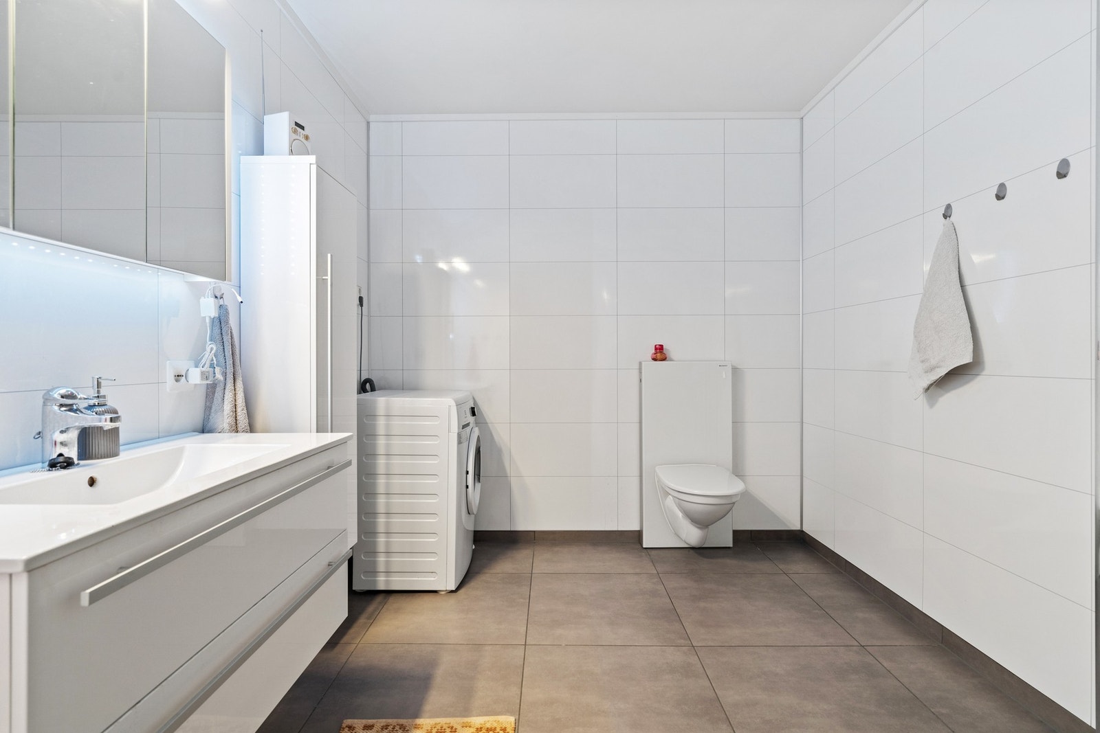 Bad/vaskerom med fliser på gulv og baderomsplater på vegger. Veggmontert toalett. Opplegg for vaskemaskin. Galleribilde