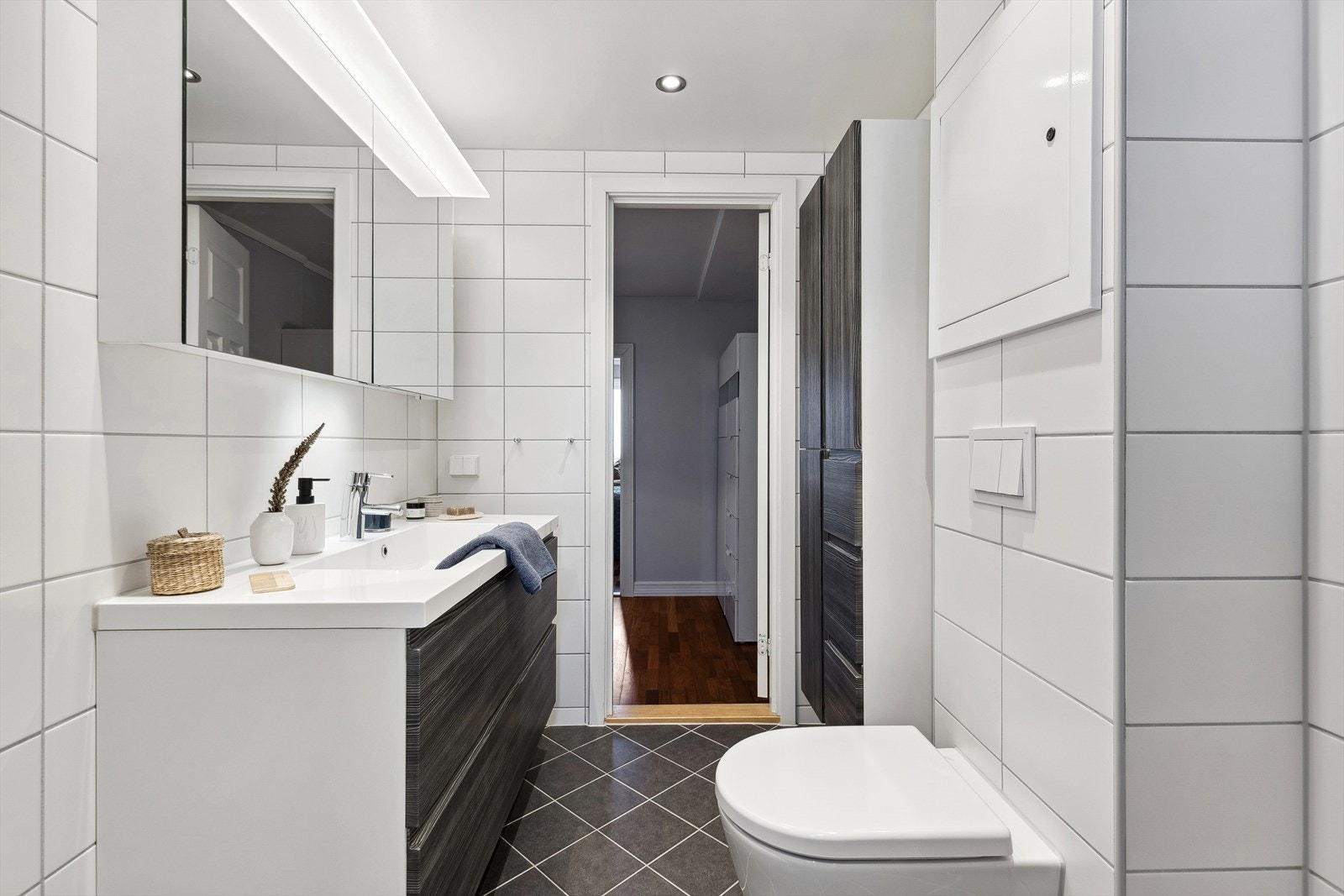 Rommet er innredet med baderomsinnredning bestående av benkeskap og høyskap med glatte fronter, veggskap med speilfronter over servant i helstøpt plate og veggfestet klosett. Galleribilde