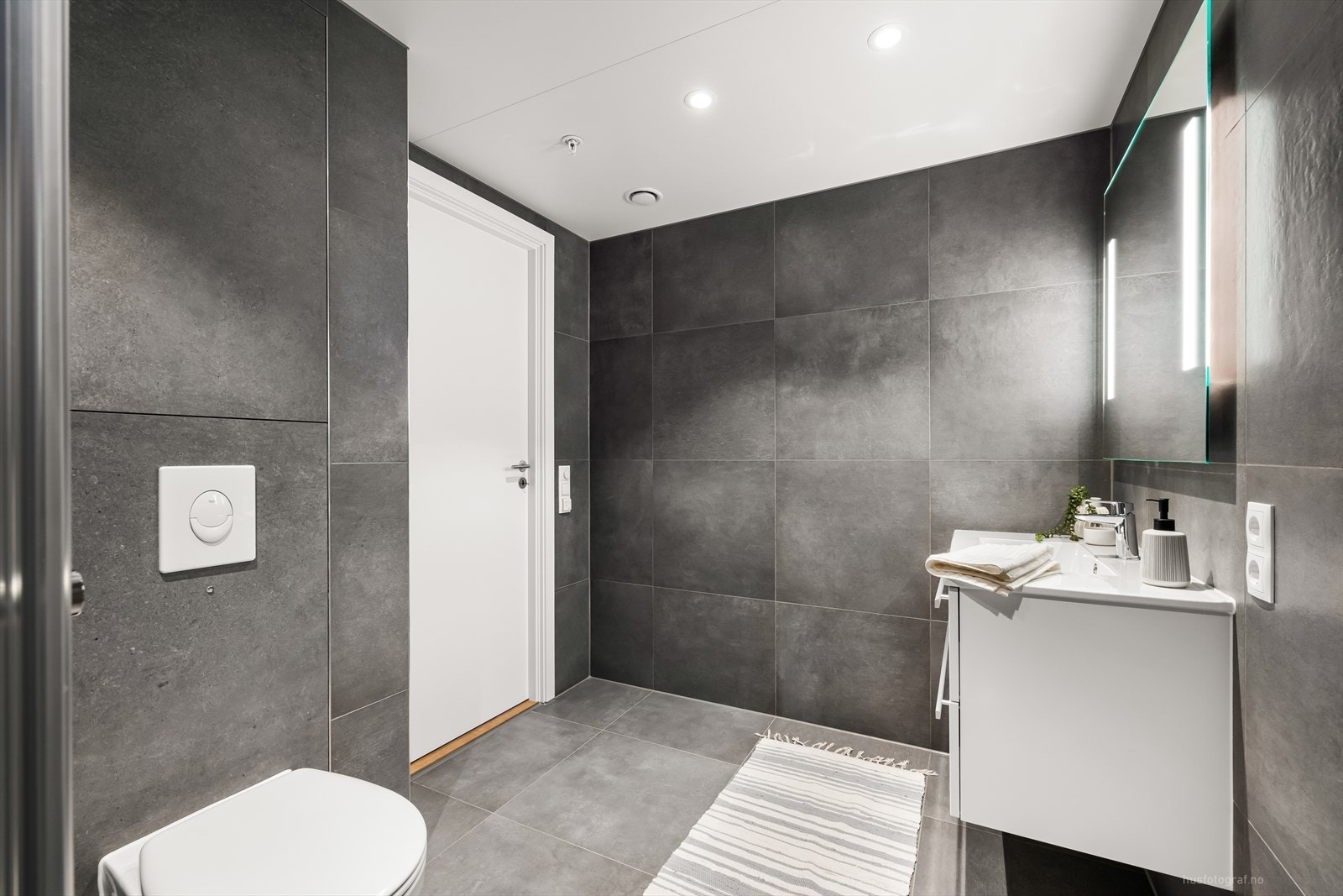 Badet består av vegghengt toalett, dusjhjørne med foldedører i glass og servant på baderomsinnredning med overhengende speil og integrert belysning. Galleribilde