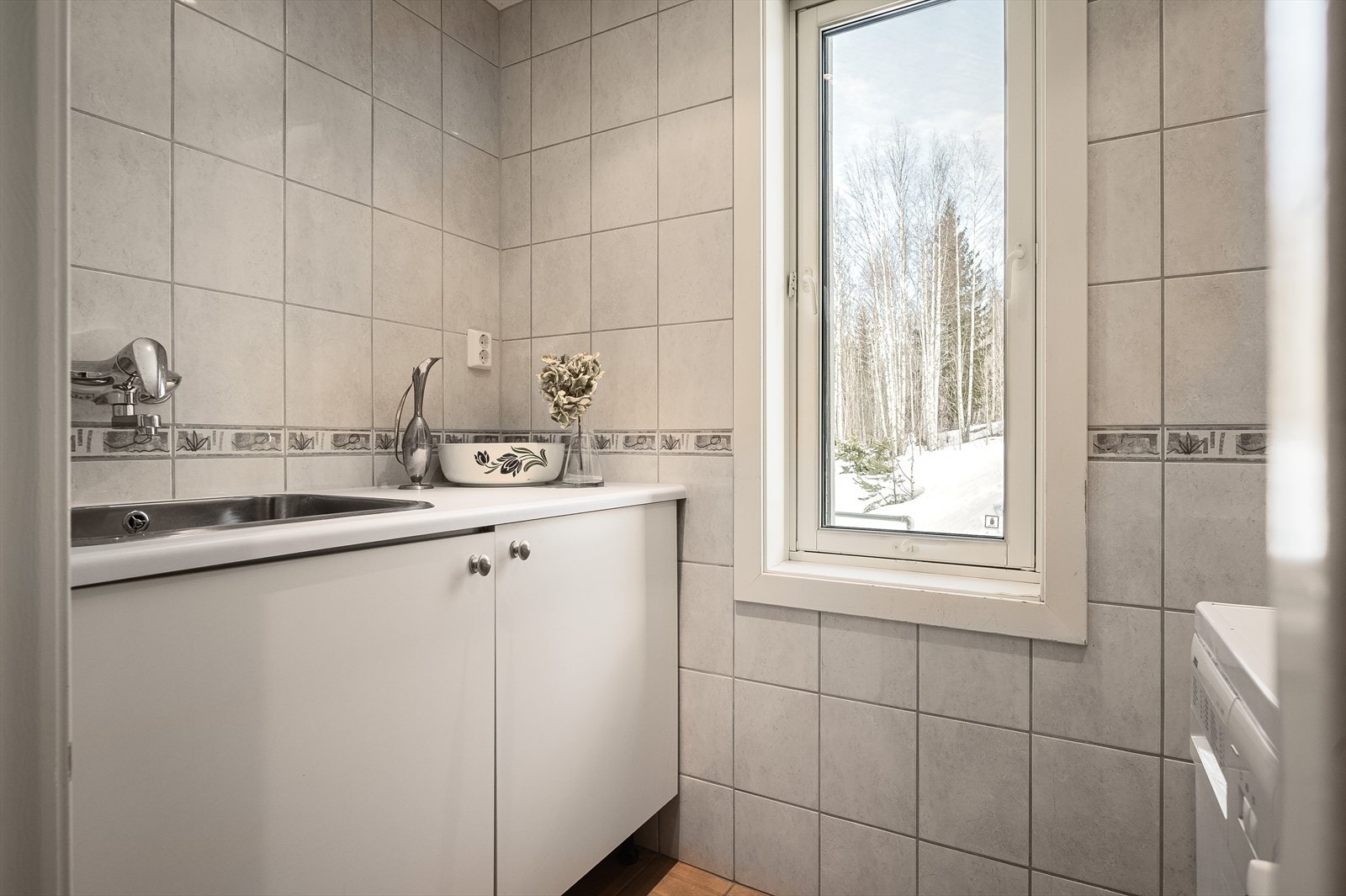 Innenfor baderommet er det et praktisk og flislagt vaskerom i samme utførelse som badet. Her er det opplegg for vaskemaskin og kondenstørketrommel, samt innredning med skap og en nedfelt servant på benkeplaten. Vindu gir fint innlys i rommet. Galleribilde