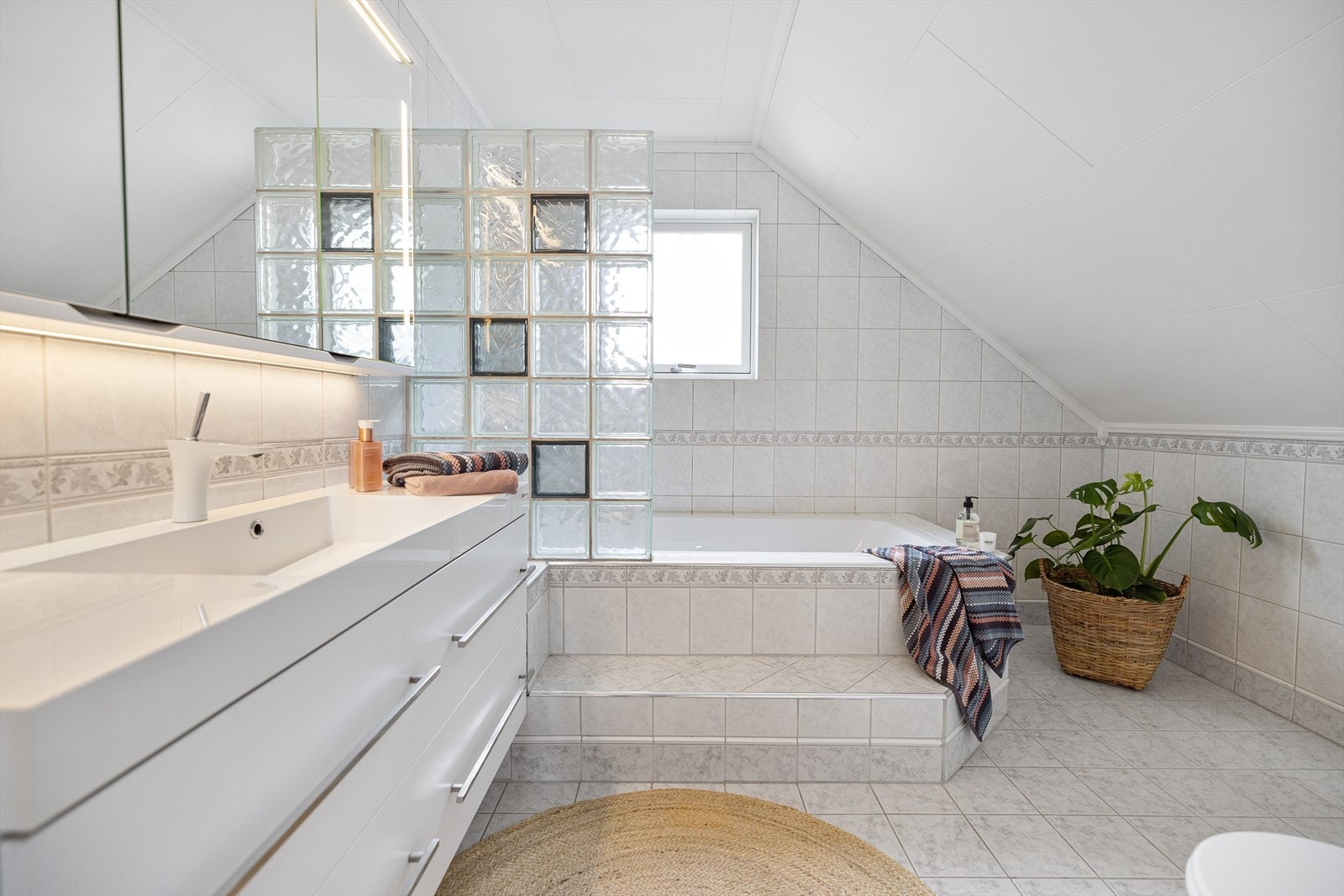 Badet er en skikkelig komfortsone - pent flislagt med en terskel opp til det innmurte badekaret som også har en dusjsone bak en vegg av glassbyggerstein. Galleribilde