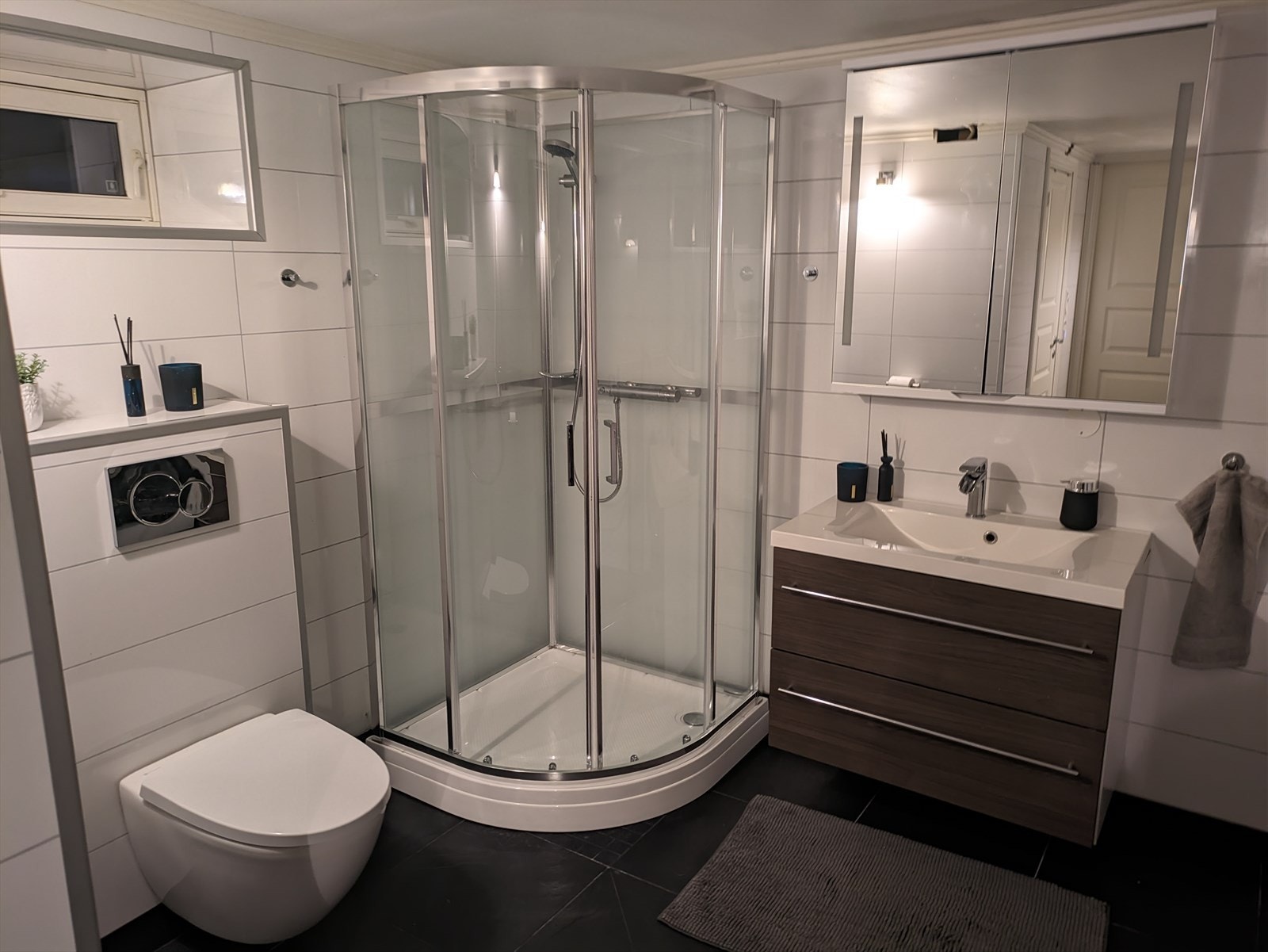 Badet har heldekkende dusjkabinett, vegghengt toalett, røropplegg for vaskemaskin og heldekkende servantplate over skuffeseksjon med glatte fronter i mørk laminat utførelse. Over servanten er det vegghengt skap med speilfronter. Galleribilde