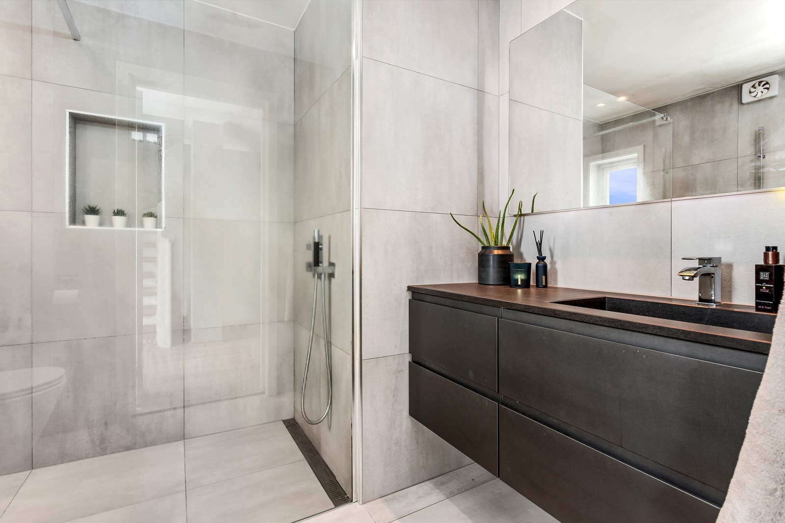 Badet har dusjplass avgrenset med glassvegg hvor det er montert dusjgarnityr med integrert røropplegg til takdusj. I tillegg er det vegghengt toalett og heldekkende servantplate over skuffeseksjoner med glatte fronter i mørklaminat materiale. Galleribilde