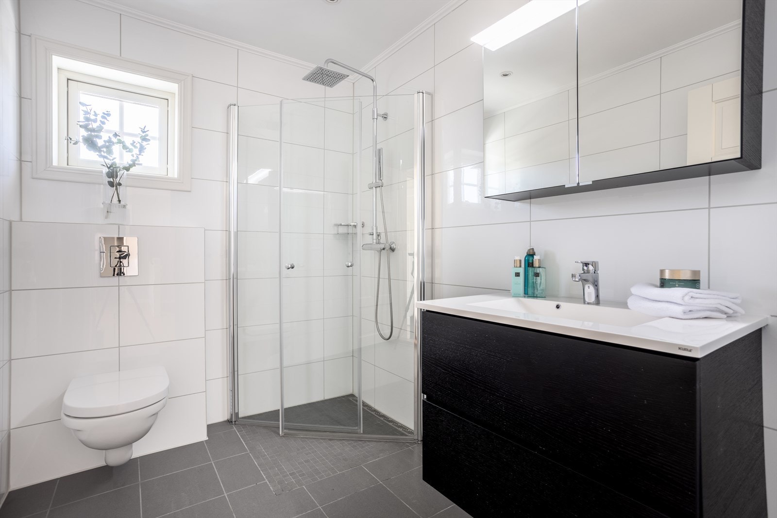 Baderom med flislagte gulv og vegger. Badet innehar vegghengt toalett, servant med skuffer og dusjnisje med innfellbare glassdører. Speilskap med belysning i overkant av servant og varmekabler i gulv. Galleribilde