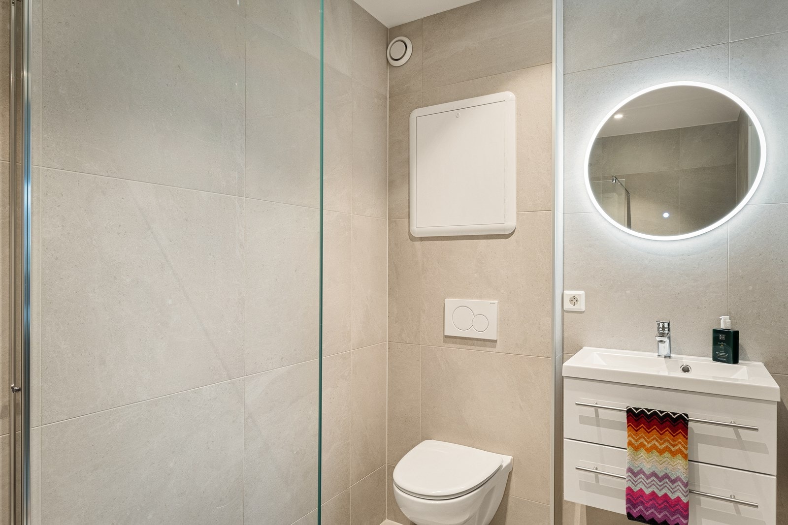 Badet inneholder vegghengt toalett, helstøpt servant, skuffer under servant med slette fronter og dusjvegg i klart glass. For ekstra komfort er bade utstyrt med varmekabler og spotter i tak. Galleribilde