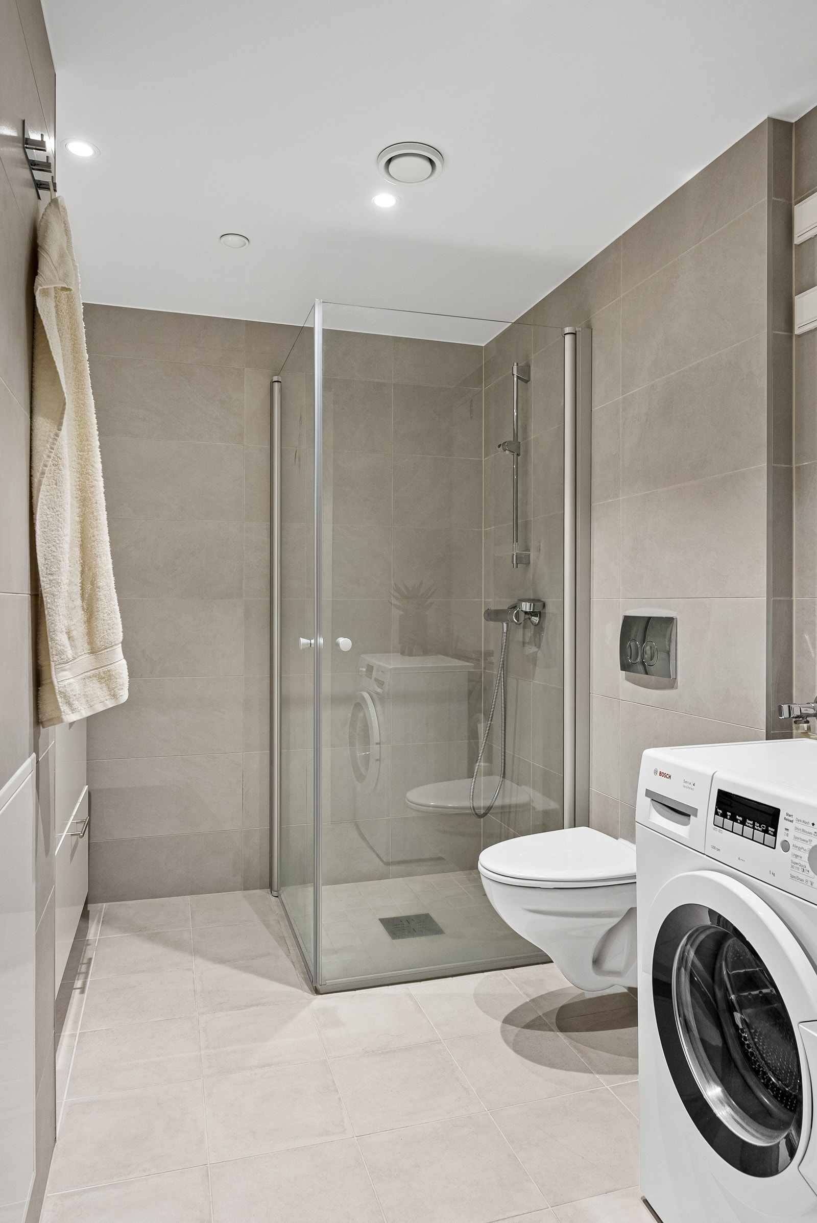 Badet har lys servantinnredning med skuffer, dusjnisje med innfellbare dusjdører i glass, veggehengt toalett, og opplegg for vaskemaskin. Galleribilde