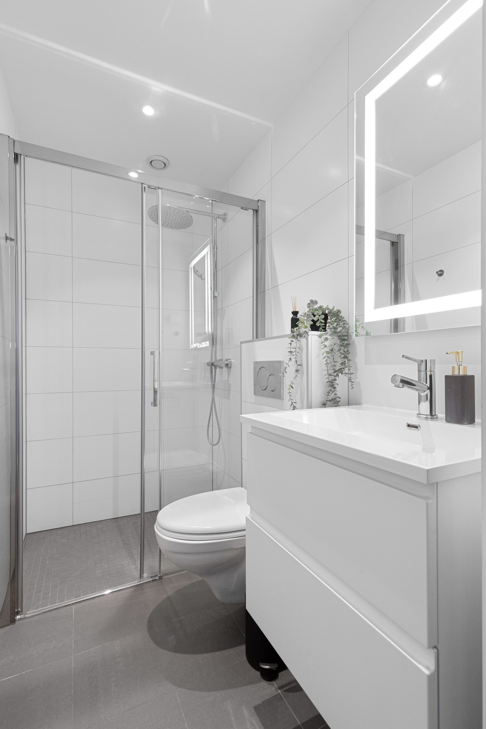 Pent, flislagt baderom som inneholder dusjhjørne, benkeskap med heldekkende servant og toalett. Galleribilde