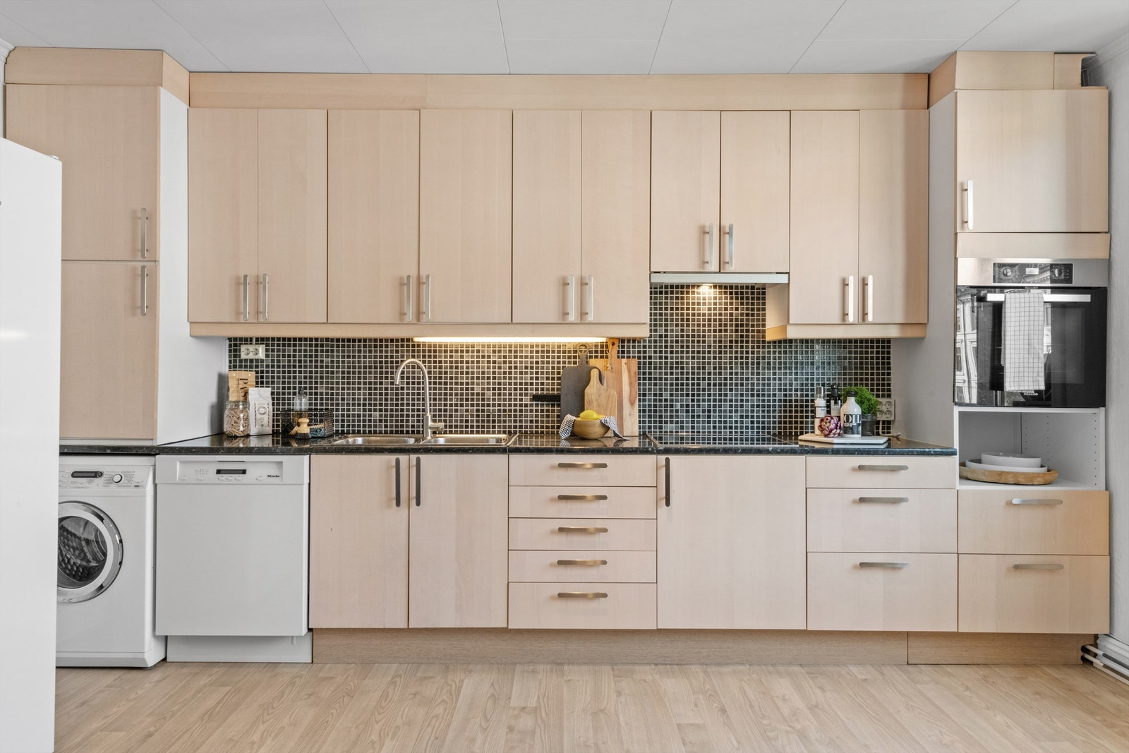 Kjøkkenet er innredet med glatte fronter, laminat benkeplate, ventilator og belysning under overskap. Induksjonstopp og stekeovn er integrert. Opplegg for oppvaskmaskin og vaskemaskin. Galleribilde