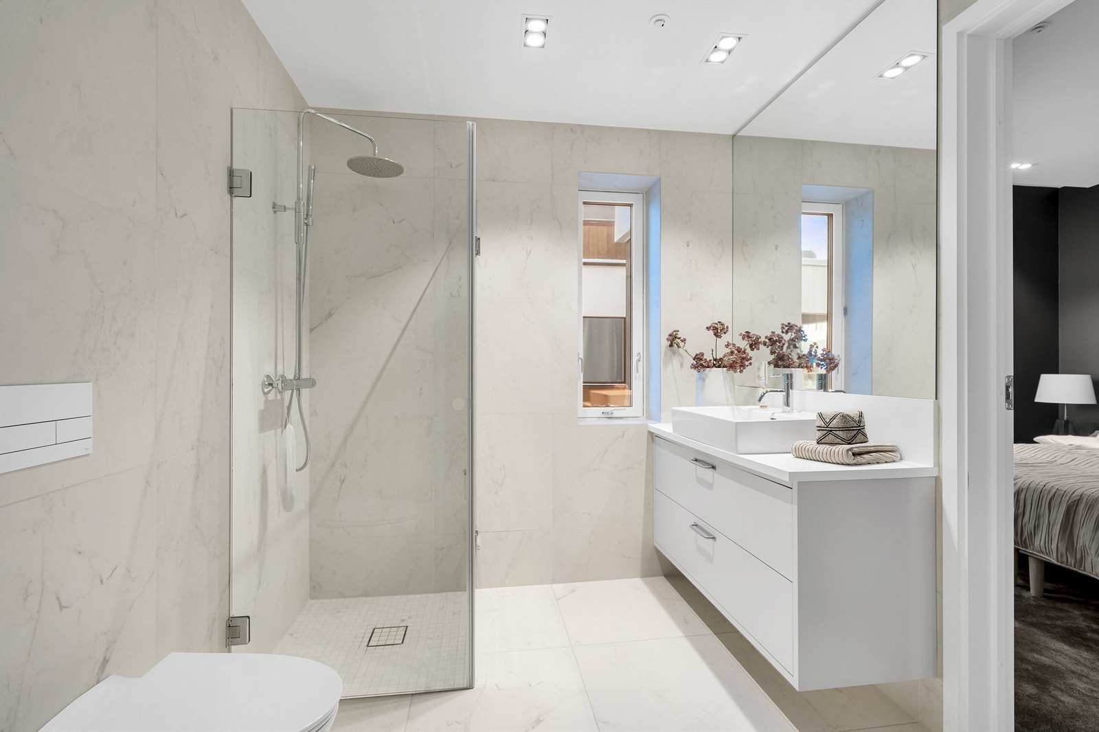 Badet er flislagt på gulv og vegger med 60x60 fliser og mosaikkfliser i dusjsonen. Det er dusjnisje med regndusj, vegghengt toalett, baderomsinnredning med servant, spotter i tak og et større speil. Galleribilde