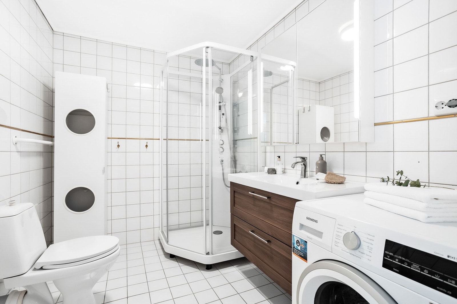 Badet har lyse fliser med varmekabler på gulv og flislagte vegger. Badet er utstyrt med servantinnredning, dusjkabinett, opplegg for vaskemaskin og wc. Galleribilde