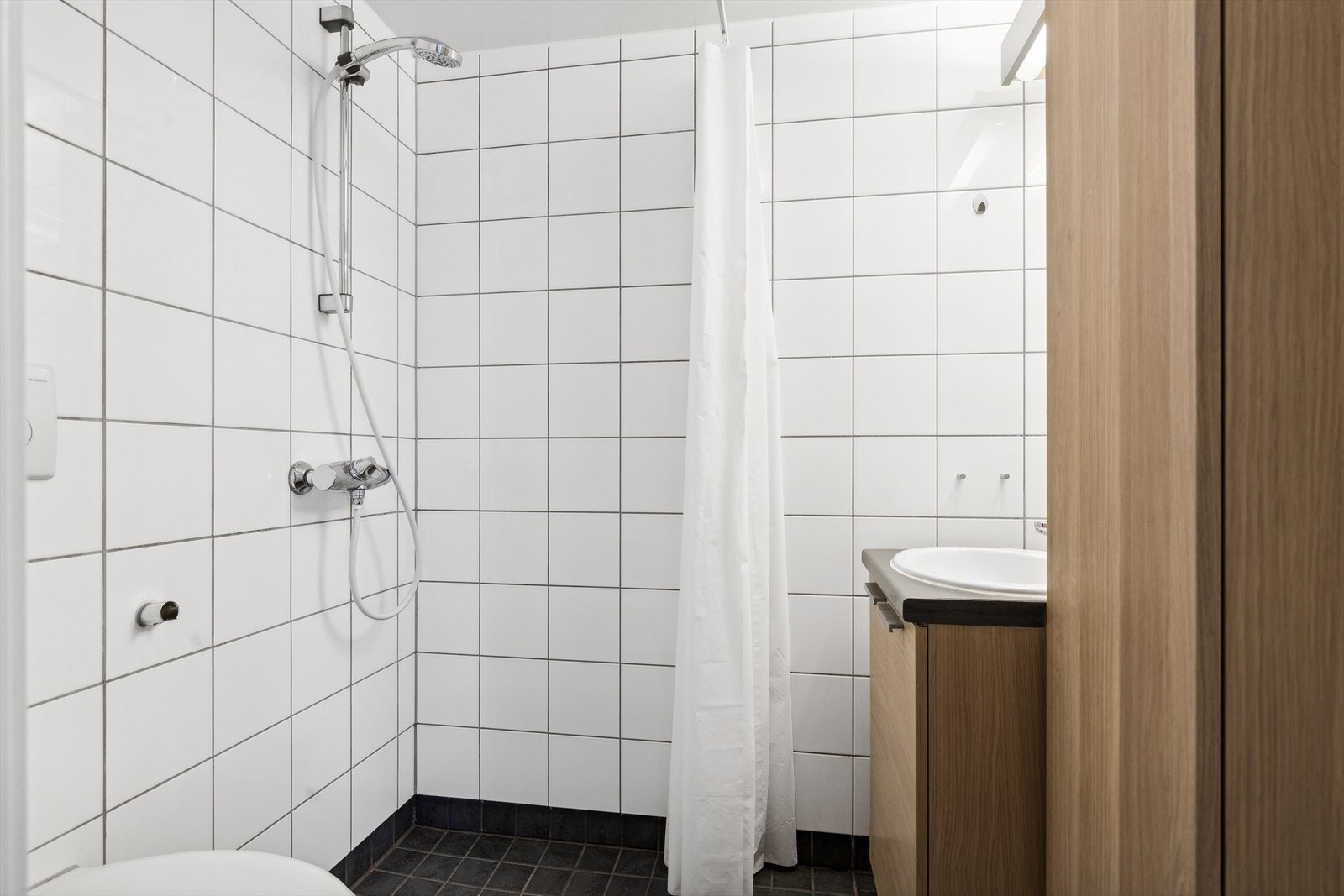 Lyst og praktisk bad med flislagte gulv og vegger. Badet er innredet med vegghengt toalett, innredning med servant og åpen dusjsone. Galleribilde