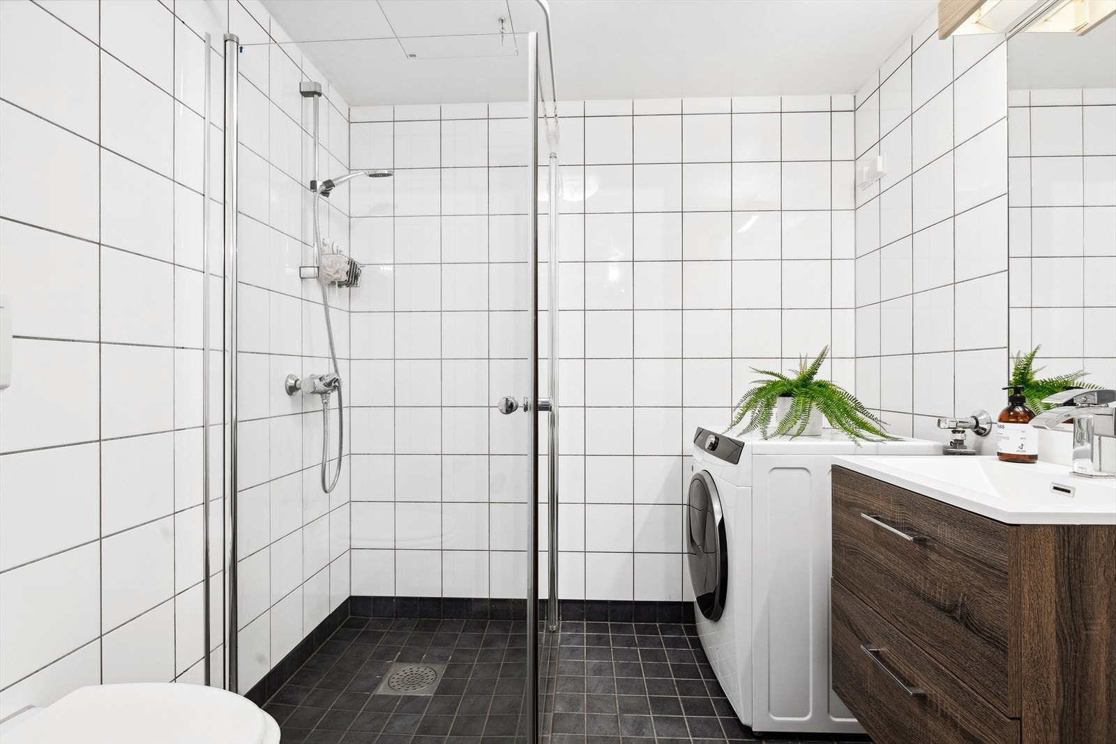 Elegant og moderne flislagt bad med opplegg for vaskemaskin. Badet er innredet med dusjvegger av glass, vegghengt toalett og servant. Galleribilde