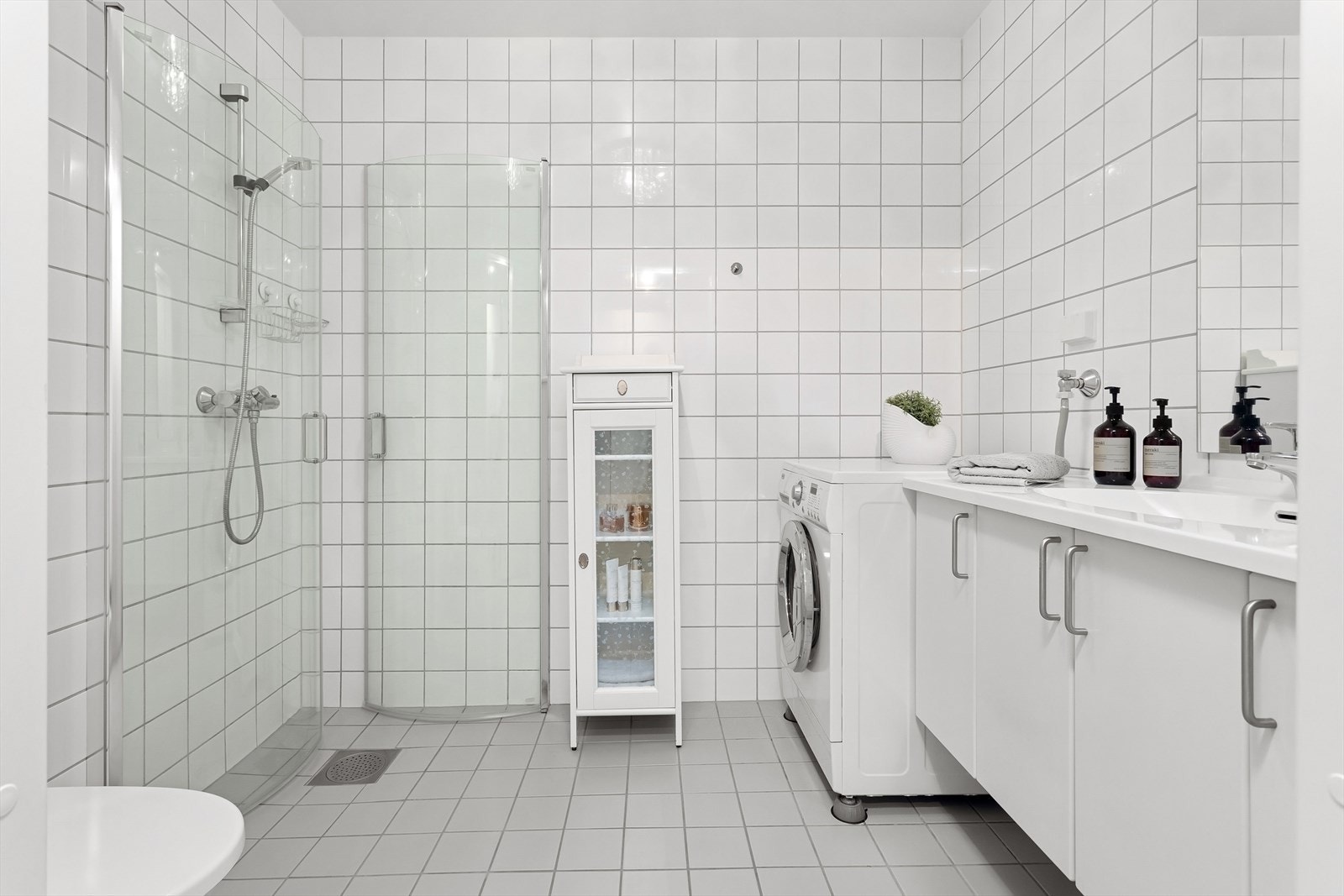 Badet har fliser på vegger og gulv. Rommet er utstyrt med WC, servant med oppbevaringsplass, dusjhjørne med innfellbare dusjdører og opplegg til vaskemaskin Galleribilde