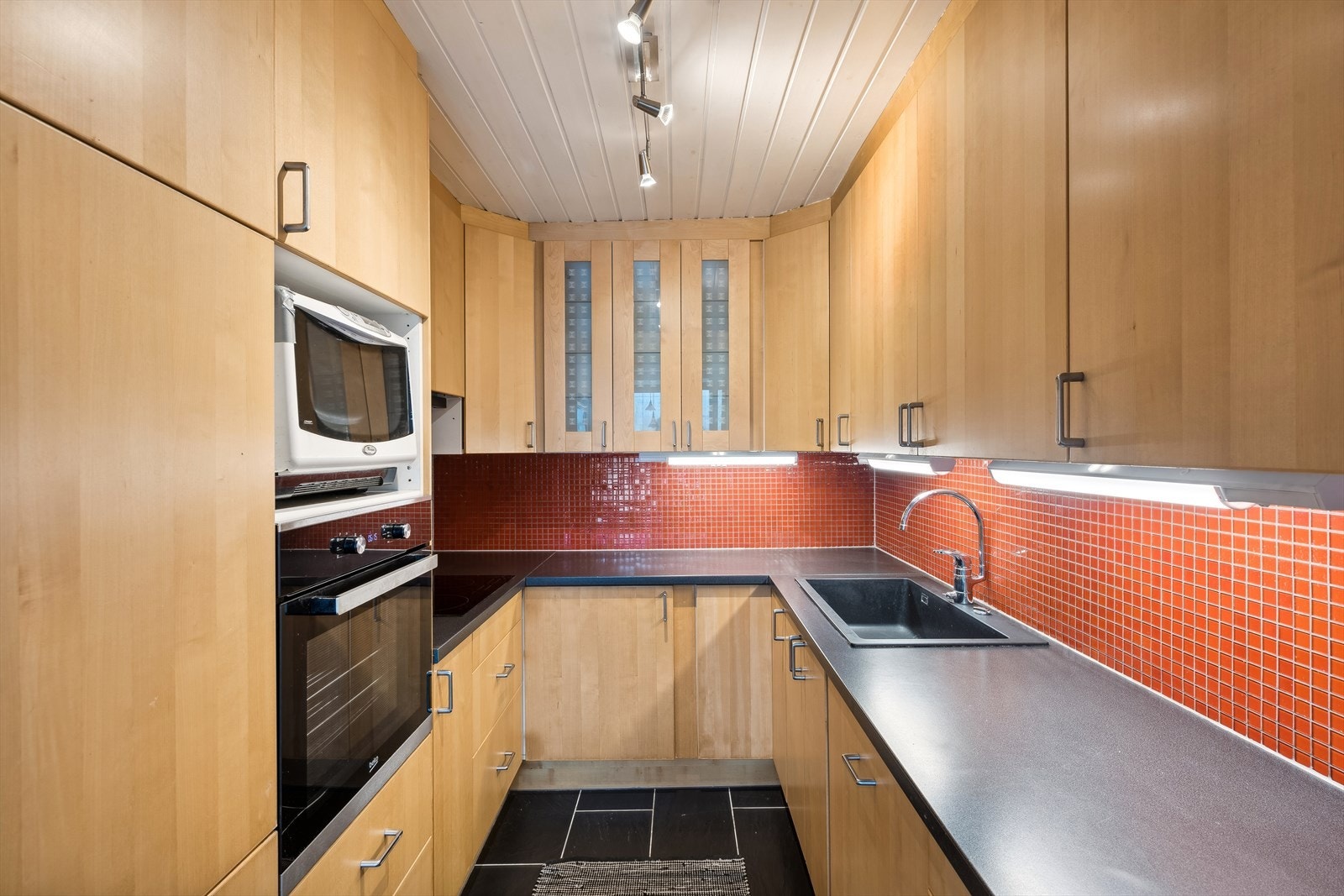 Det er glatte trefargede fronter og benkeplate i laminat med oppvaskkum i kompositt. Over benkeplaten er det belysning og dekorative fliser som gjør rengjøringen enklere. Galleribilde