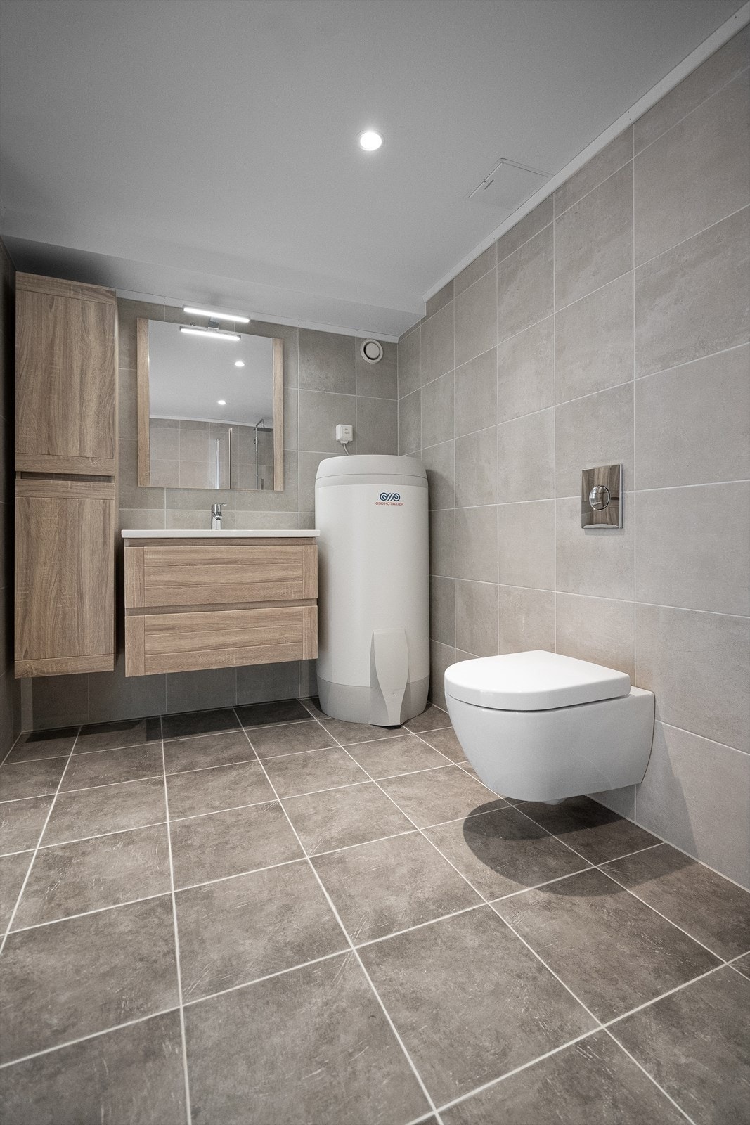 Moderne baderomsinnredning med servant, speil med belysning over og vegghengt skap ved siden av. Baderommet er også utstyrt med dusjhjørne med glassdører og regnfallsdusj og vegghengt toalett. Galleribilde