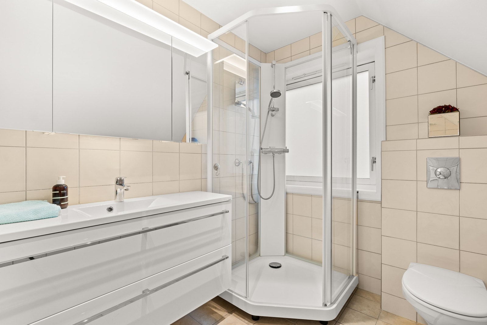 Baderommet har fliser på vegger og gulv, samt gulvvarme, og er utrustet med vegghengt servantinnredning med heldekkende servant, speilskap med belysning og stikkontakter montert over servant, dusjkabinett med skyvedør av glass, samt vegghengt toalett Galleribilde