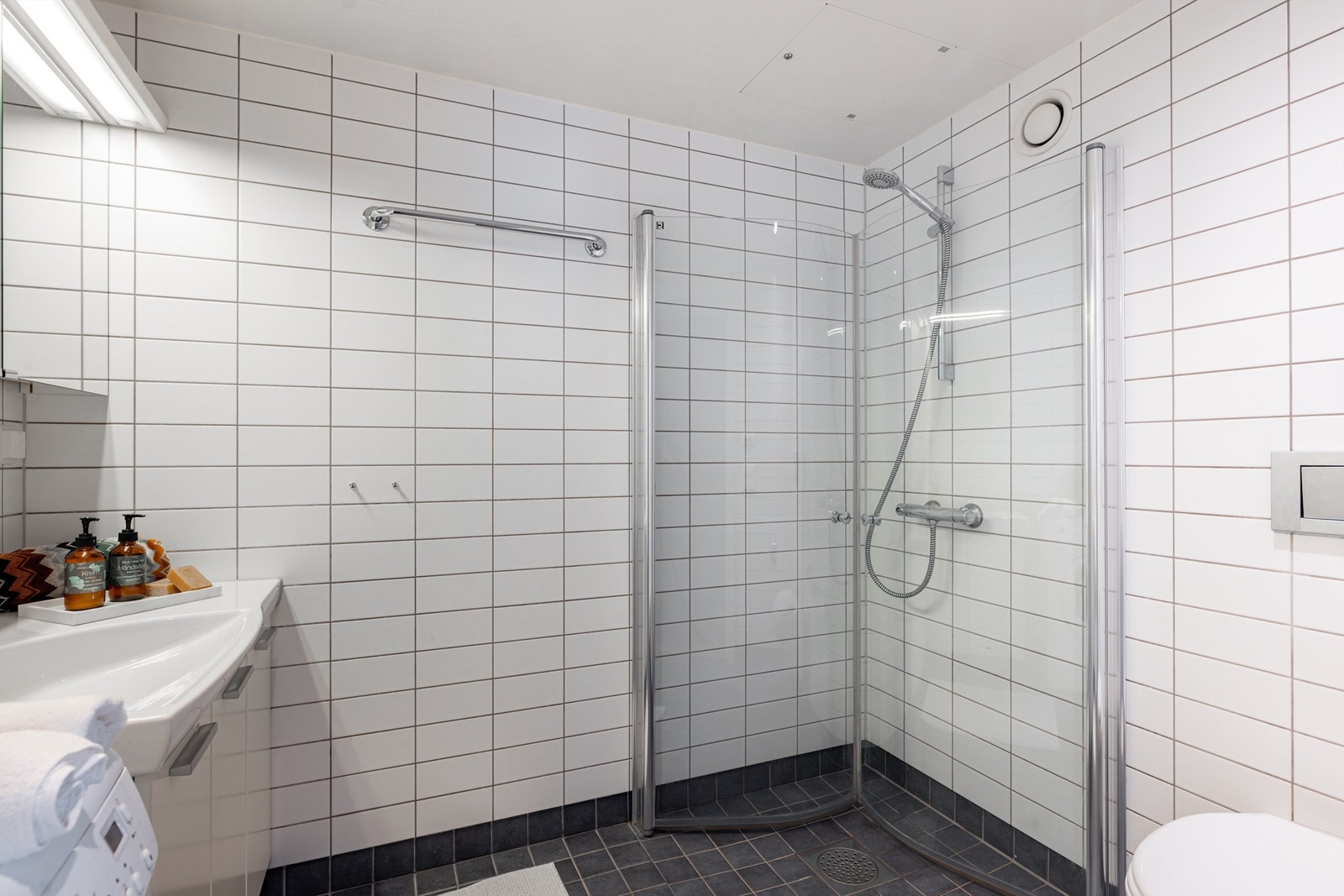 Badet har dusjhjørne med foldedører av glass, vegghengt toalett og opplegg for vaskemaskin Galleribilde