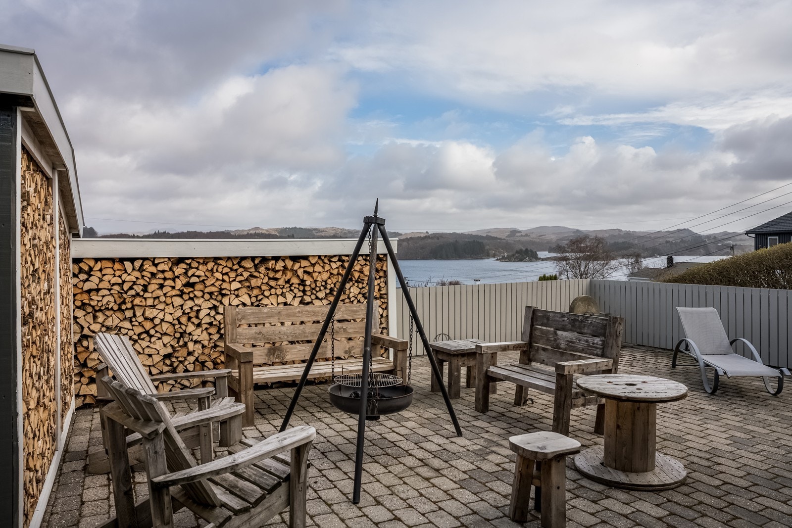 Nederste terrasseområde med fint lagt belegningsstein, lave rekkverk mot fjordutsikten (for å nyte utsikten fra sittestilling i nederste hagestue). Kjekk bålpanneplass med plass for stor stittegruppe. Stort takoverbygg for ly mot regn. Galleribilde