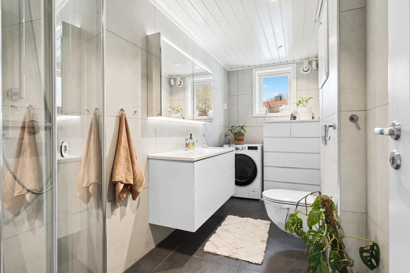 Baderommet har dusjvegger av glass, vegghengt toalett, gulvmontert toalett og opplegg for vaskemaskin. Galleribilde