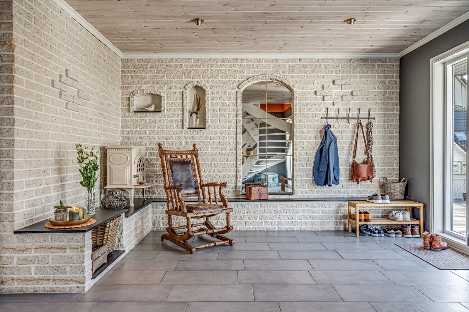 Inngang til en svært romslig hall med fliser på gulv, trapp opp til loftsetasjen, fint integrert speil i murvegg, vedovn, spotter i tak og vinduer nesten fra gulv til tak. Galleribilde