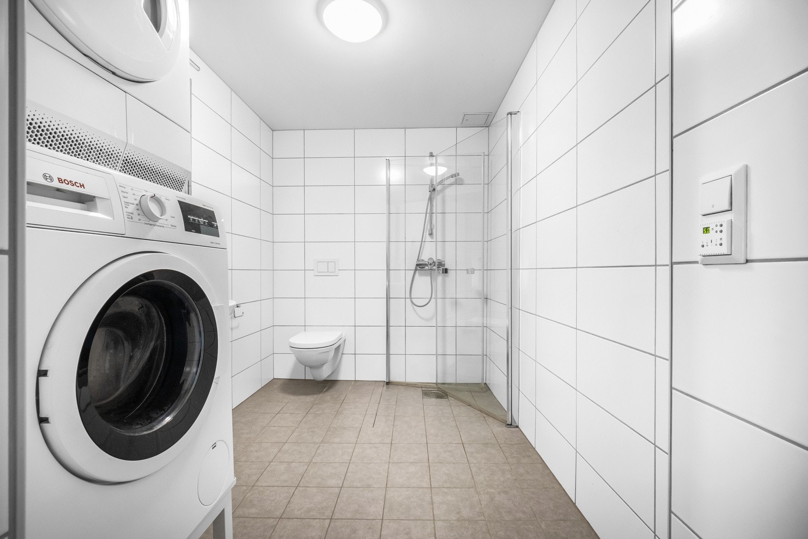 Tidløst flisvalg med grå gulvfliser og hvite veggfliser. Stort dusjhjørne med foldbare glassdører, vegghengt wc og hvit servant med speilskap. Galleribilde