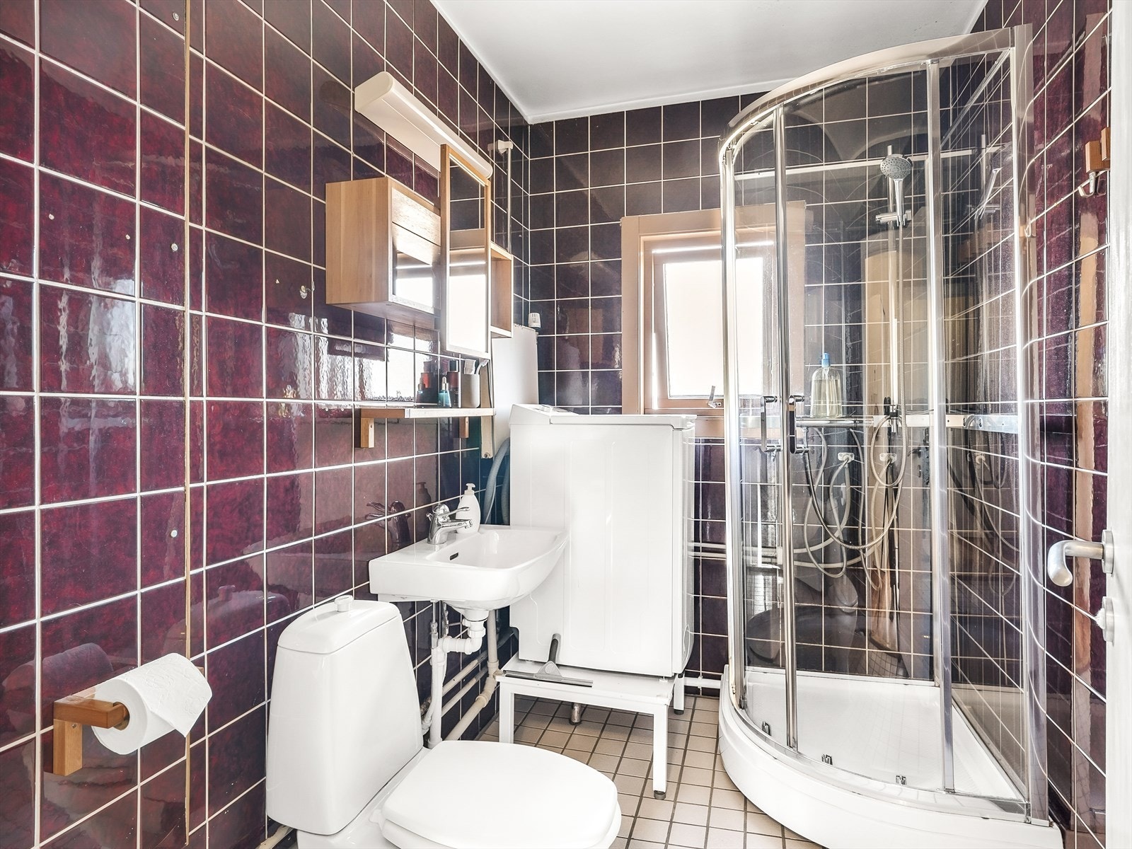 Bad med fliser på gulv og vegger samt malt tak. Innredningen består av wc, dusjkabinett, servant, speilhylle. veggskap og opplegg for vaskemaskin. Galleribilde