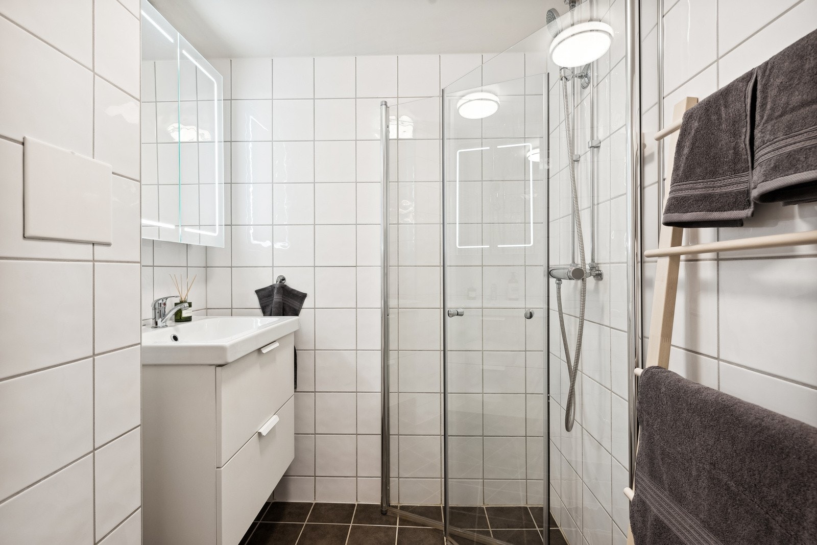 Badet inneholder vegghengt servantskap, ovenpåliggende servant med ett-greps armatur, speilskap med belysning og stikkontakt på vegg over servant. Dusjsone med innfellbare dører av glass, dusjarmatur tilkoblet hånddusj, gulvstående toalett. Galleribilde