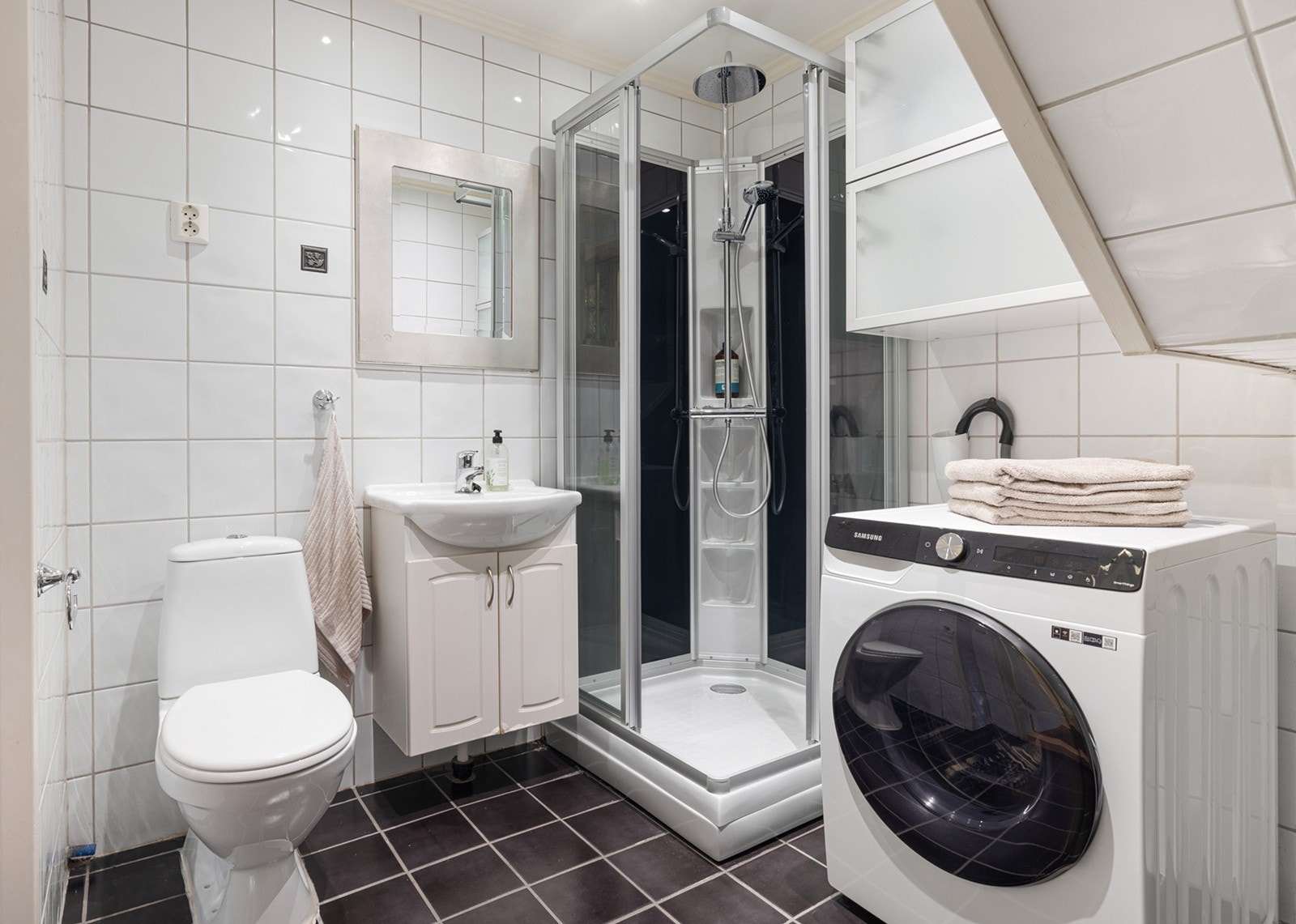Bad med hvite veggfliser, og sorte gulvfliser med gulvvarme. Innredningen består av servant med underskap, speil og stikk. I tillegg har badet et dusjkabinett med regnfallsdusj, hånddusj og glassdør.
Det er opplegg for vaskemaskin. Galleribilde