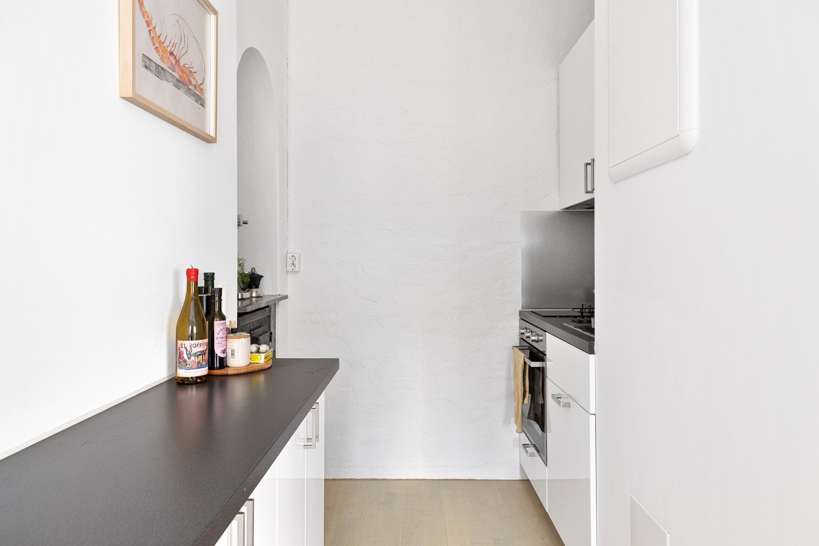 Hybelkjøkken med hvite høyglansfronter og stålhåndtak, overskap og børstede stålplater på vegg Galleribilde