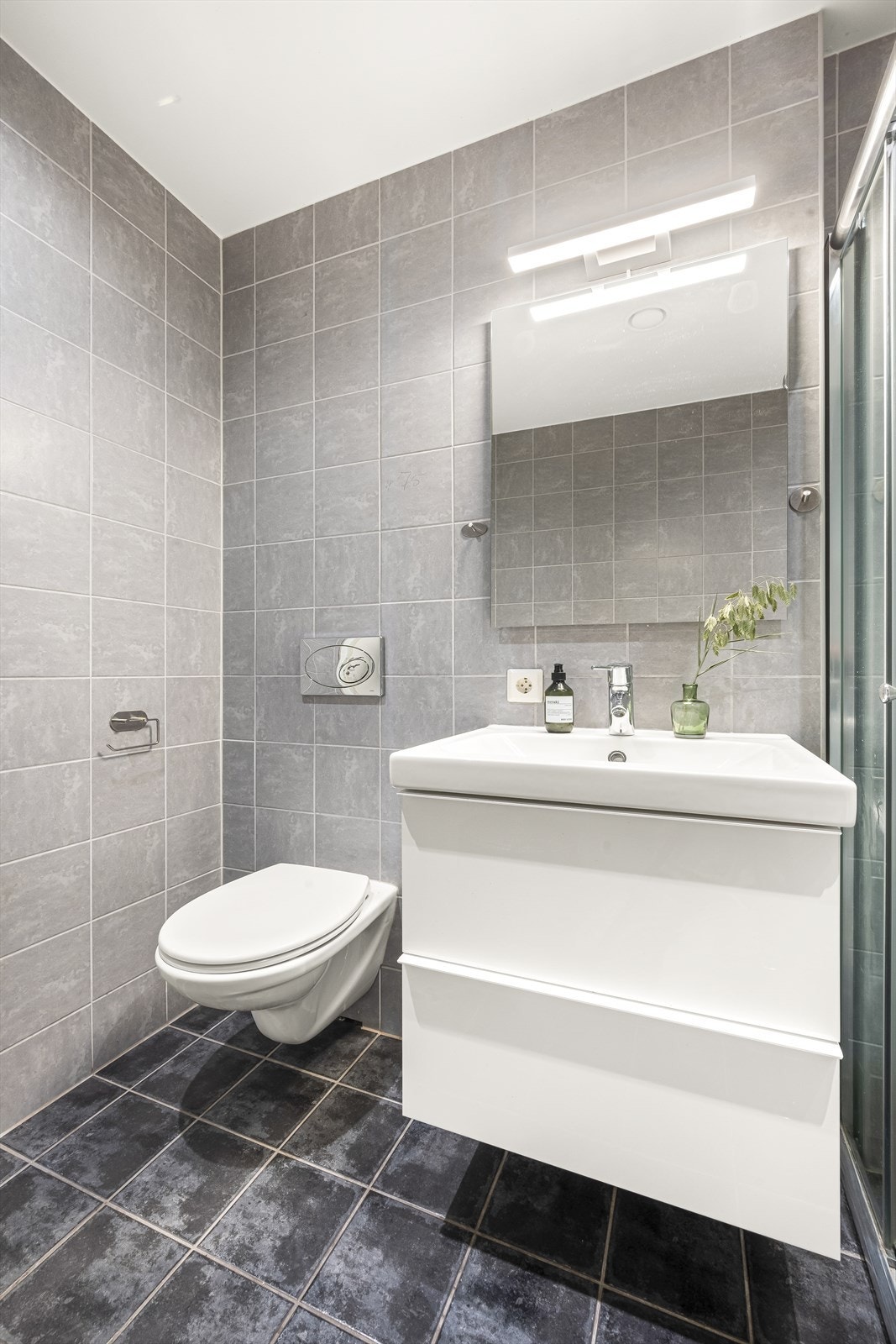 Baderomsinnredningen inneholder videre underskap, servant, speil, stikk og nedsenket dusjsone med dusjing direkte på flisene. Videre er det tilkoblet et klosett og opplegg for vaskemaskin. Badet har balansert ventilasjon. Galleribilde