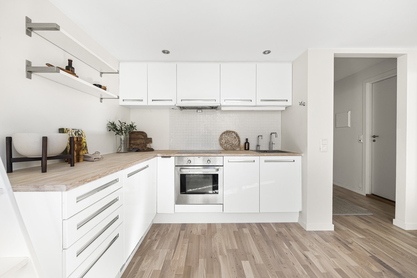 Kjøkkeninnredning med bambus benkeplate, hvite slette fronter og fliser mellom over og underskap. Det er montert en integrert oppvaskmaskin, komfyr, kjøleskap, nedfelt platetopp og det er tilkoblet en ventilator over kokeplass. Galleribilde