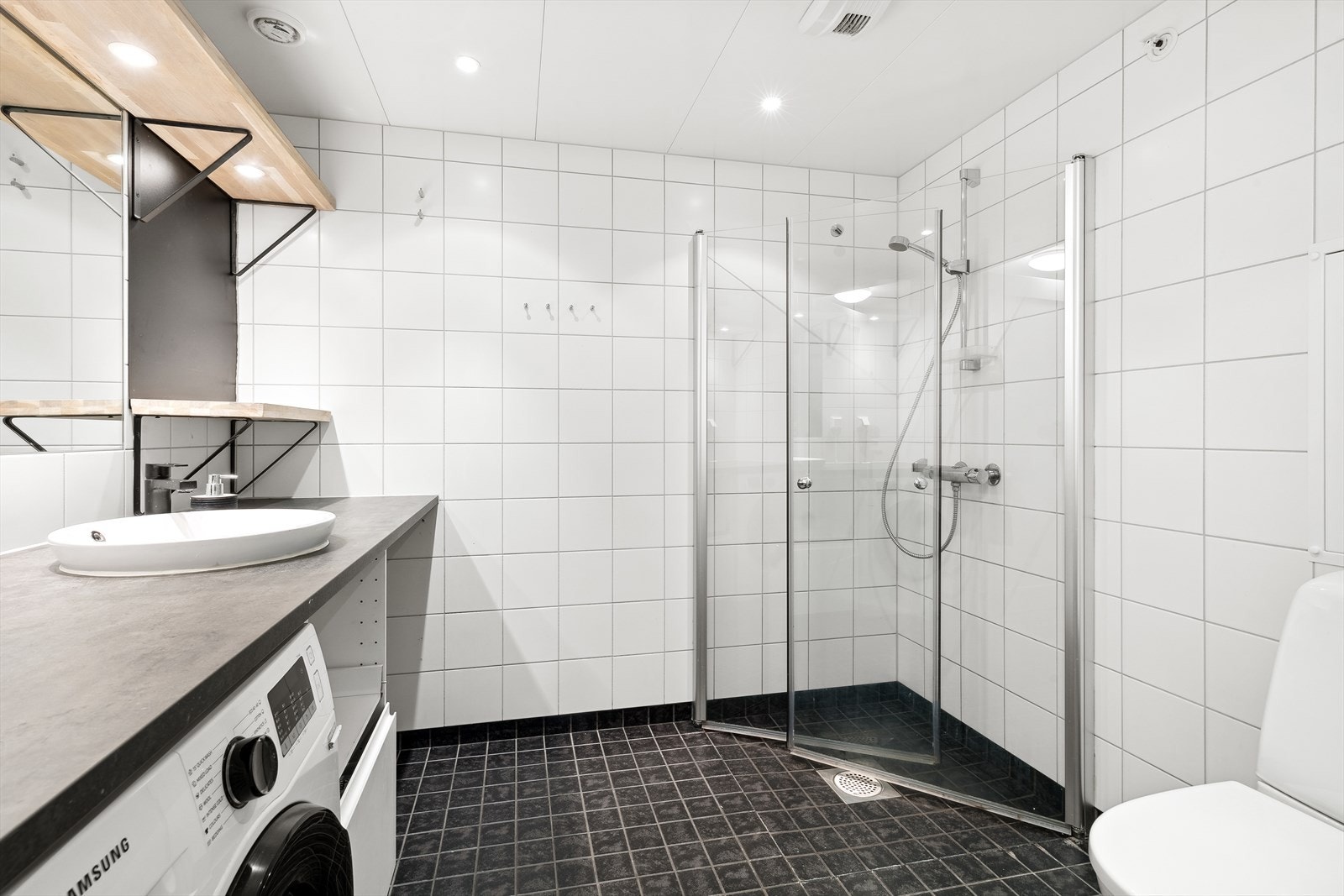 Romslig og pent flislagt bad med vannbåren gulvvarme og downlights i himlingen. Badet er utstyrt med dusjhjørne med innfellbare glassvegger, vegghengt wc, servantinnredning med skuff, samt opplegg og plass til vaskemaskin under praktisk brettebenk Galleribilde