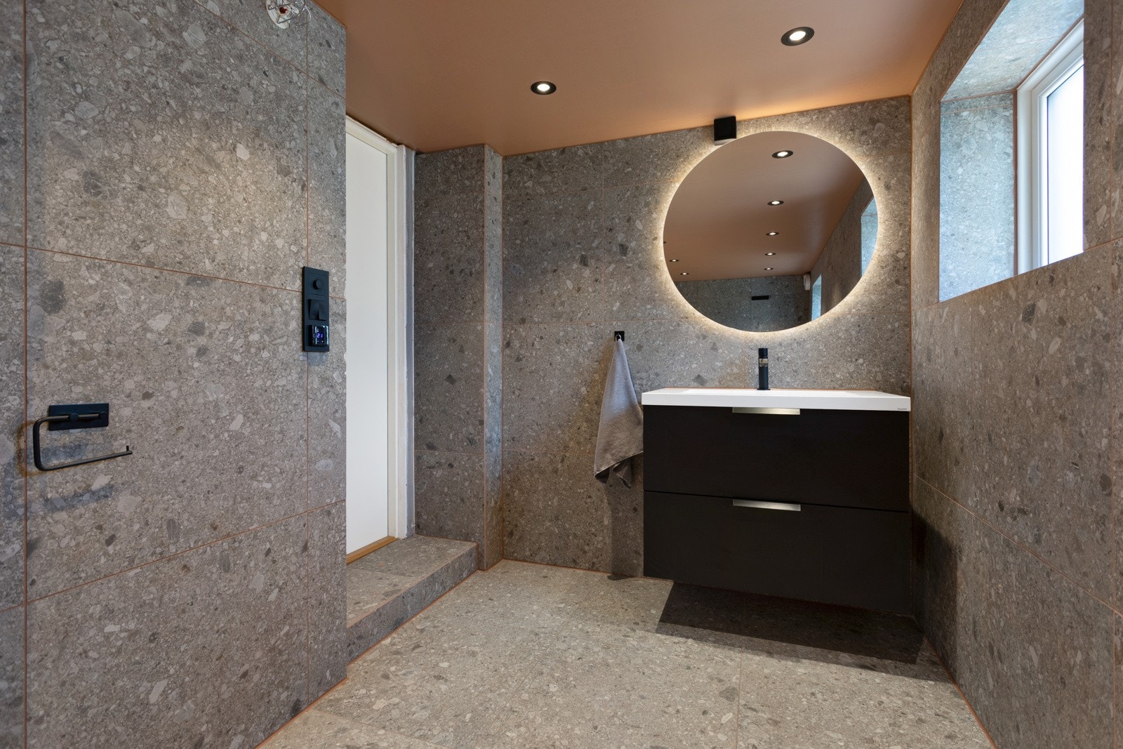 Det ene badet har betonggrå fliser i steinmønster på gulv og vegger, vinduer og downlights. Taket er malt orange. Rommet er pent innredet med heldekkende servant på sort skuffeseksjon, rundt speil med belysning, veggmontert toalett og et dypt badekar. Galleribilde