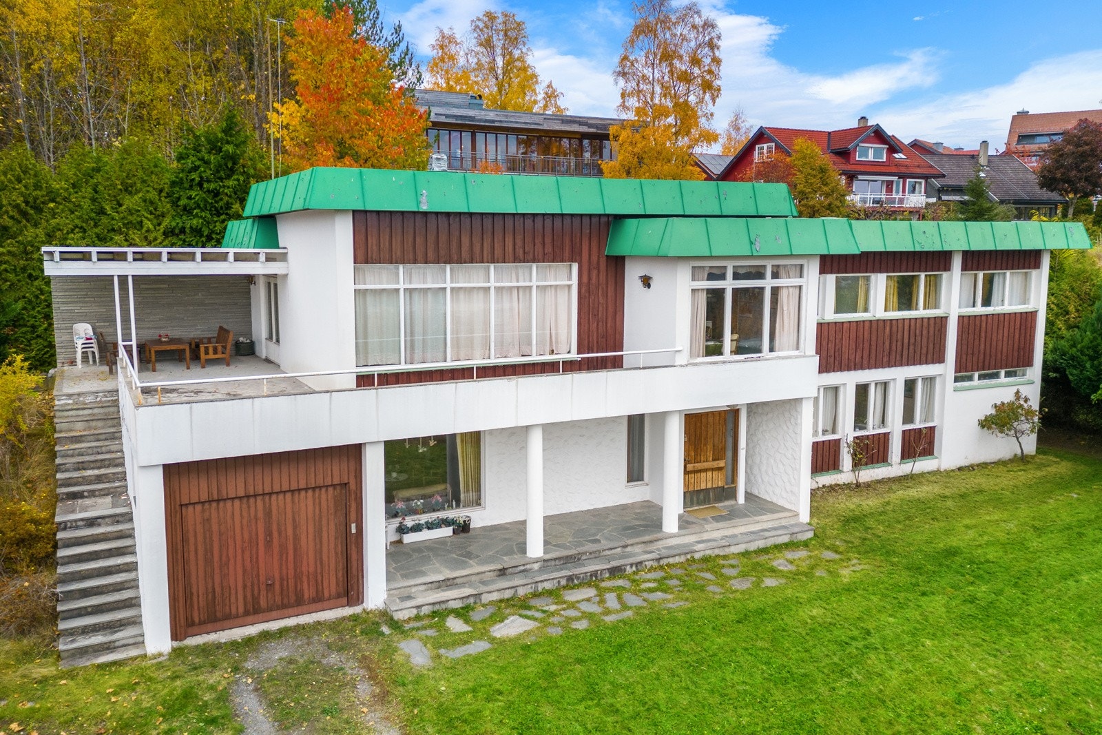 Fasadebilde som viser inngangsparti, store vinduer som vender mot syd, stor terrasse og med garasje i underetasjen Galleribilde