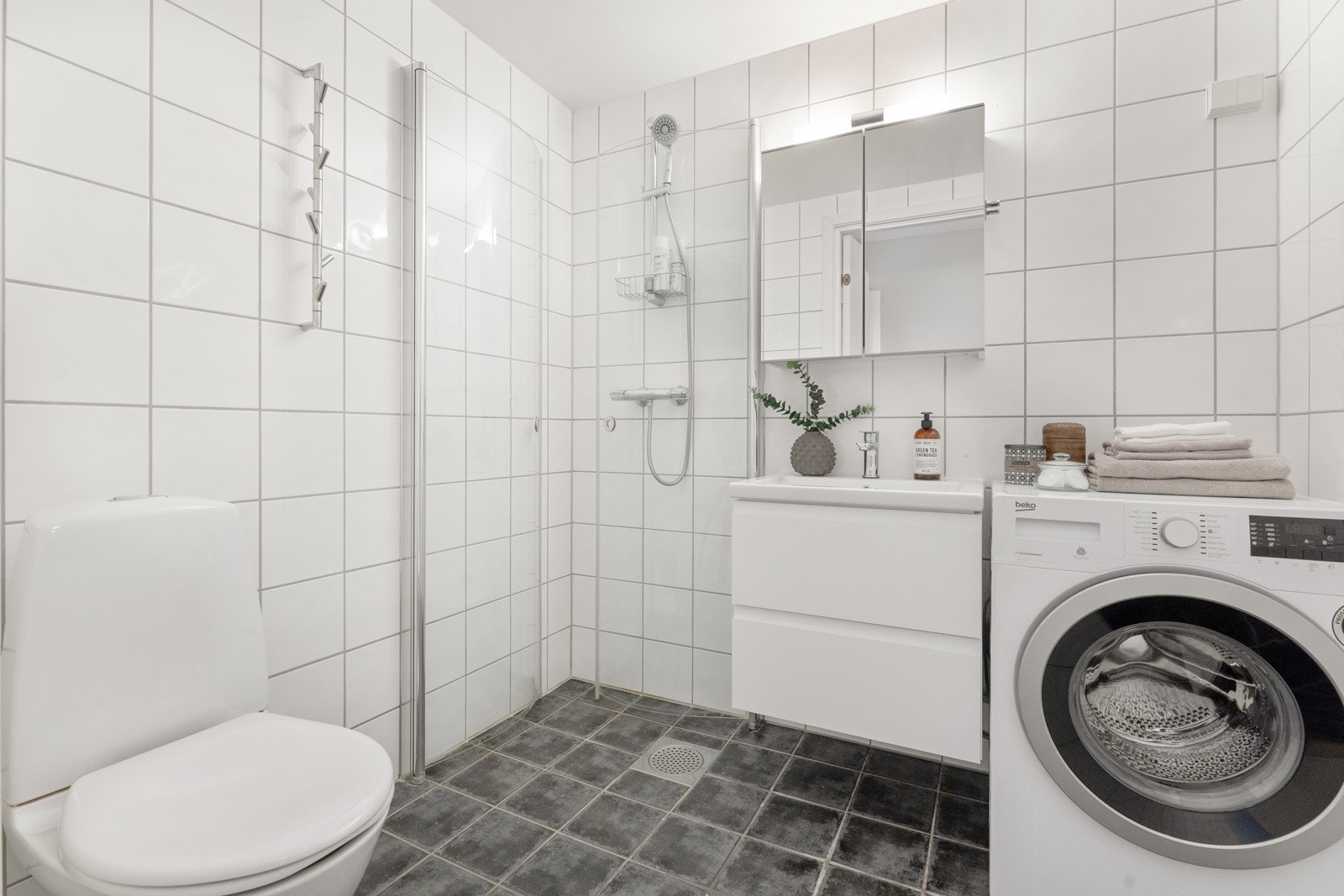 Badet er delikat med mørkegrå gulvfliser, hvite veggfliser, gulvvarme og mekanisk avtrekk. Badet har toalett, dusjhjørne med buede, innfellbare fronter og opplegg for vaskemaskin. Galleribilde