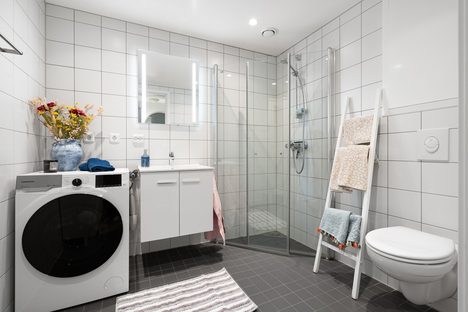 Lyst og tidløst flislagt bad med varmekabler i gulv, innfellbare dusjdører i glass og opplegg til vaskemaskin Galleribilde